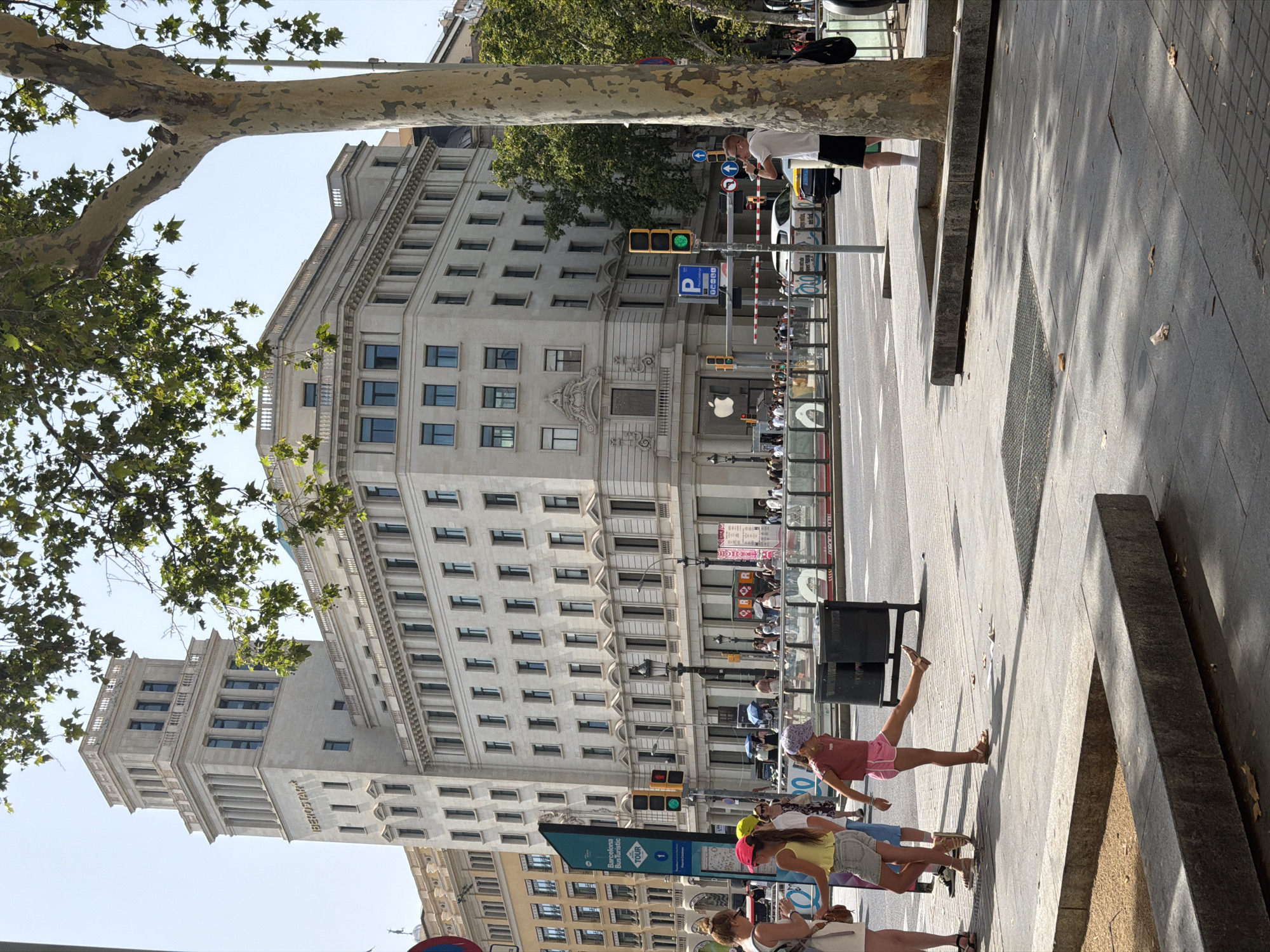The image depicts a sunny street scene in Barcelona, Spain, likely on a warm day, as indicated by the presence of people in light clothing and the shadows cast by trees. In the background, a large, ornate building, which appears to be a hotel or a significant commercial establishment, dominates the skyline. The building has a distinctive architectural style, with a prominent tower featuring classical columns and a modernistic element at its base, where "IBEROSTAR" is inscribed.
In the foreground, a young girl in a pink t-shirt and shorts is captured mid-action, kicking her right leg out. She is wearing sandals and a patterned cap. To her left, another young girl in a yellow top and denim shorts is standing. A woman in a white dress is also visible to the far left. They are standing on a sidewalk, next to a large tree with textured bark.
Across the street, traffic lights are visible, with one showing a green light. A sign with a blue background and white text, possibly indicating a bus route or tourist information, is present. There are also several trash bins on the sidewalk. Further down the street, a white car is parked, and a man in a white t-shirt and black shorts is standing near a tree, seemingly taking a photograph or recording with his phone. The overall environment suggests a busy urban setting with pedestrian traffic and vehicular movement. The architecture, signage, and overall atmosphere are consistent with a location in a major European city like Barcelona. The presence of an Apple Store logo on the building in the background further narrows down the potential area to a prominent commercial district within the city.