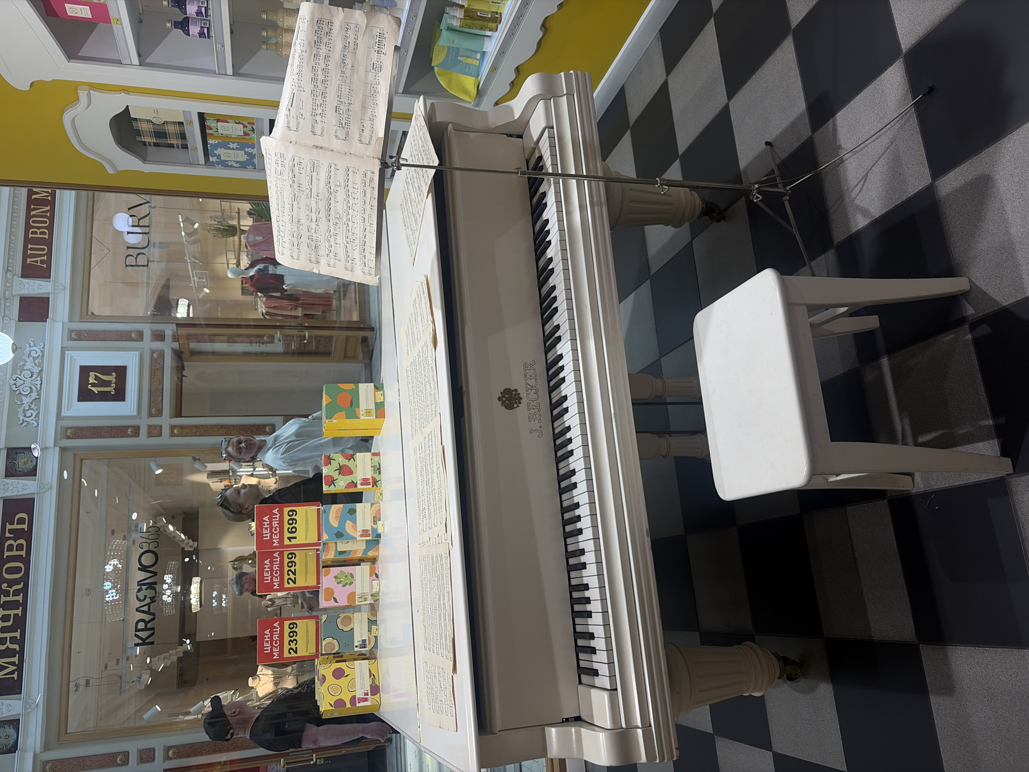 A white grand piano with sheet music on a stand is placed in front of a store window displaying various products. Three price tags with Cyrillic text and numbers are visible in the window, with prices of 2399, 2299, and 1699. Several colorful boxes are arranged in front of the price tags. In the background, mannequins are displayed in a store. The floor is a black and white checkered pattern. The piano has the inscription "J. BECKER" on it. A white stool is positioned to the right of the piano. This scene is located in Saint Petersburg, Russia.