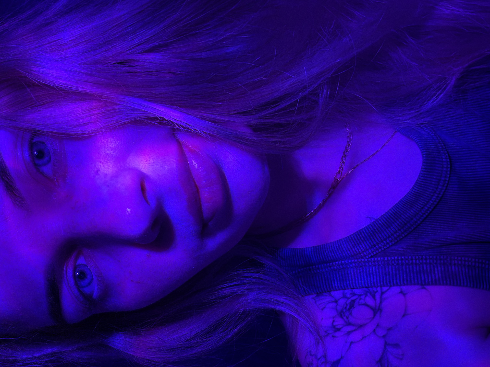 A close-up photograph features a woman with light blonde hair illuminated by a strong blue light. The woman is lying down, angled towards the camera. Her facial features are partially obscured by the blue light, but her eyes are visible and appear light-colored. She has a gold chain necklace around her neck. Part of her torso is visible, covered by a dark ribbed top. A floral tattoo is visible on her chest or shoulder area. The overall setting is dark, with the blue light highlighting the woman's features. The location, Chaikovsky, Russia, is not discernible from the image.