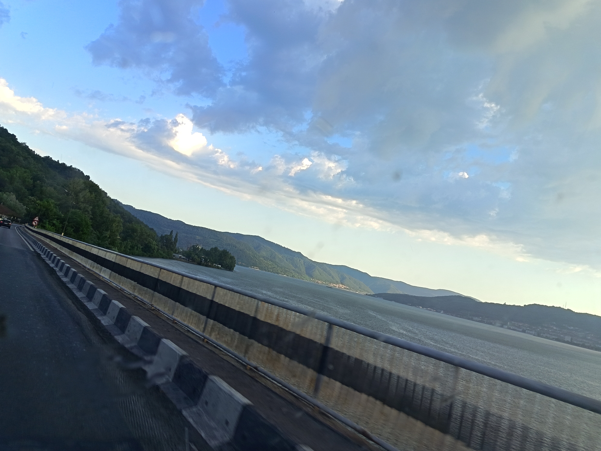 The media file captures a scenic view from a bridge, likely on a highway, overlooking a wide river or body of water. The foreground is dominated by the road and a concrete barrier with black and white stripes, indicative of a highway safety measure. The road surface appears wet, suggesting recent rain or mist. On the left side of the road, a lush green, tree-covered hillside slopes down towards the water. A few dark-colored vehicles are visible on the road, moving away from the viewer.
The main subject of the image is the expansive body of water, which stretches out to the horizon. The water is calm, with gentle ripples reflecting the sky. Across the water, a series of rolling, green hills form a dramatic backdrop. Some structures, possibly buildings or a town, are subtly visible on the far shore, nestled amongst the hills.
The sky above is a dynamic mix of blue and white clouds. Some clouds are bright and puffy, catching the sunlight, while others are more diffuse and grey, creating a sense of depth and movement. The lighting suggests it is either early morning or late afternoon, with a soft, natural light illuminating the landscape.
The overall mood is peaceful and serene, evoking a sense of travel and natural beauty. The location, identified as Orșova, Romania, places this scene along the Danube River, a significant waterway in the region. There is no visible text in the media file.
