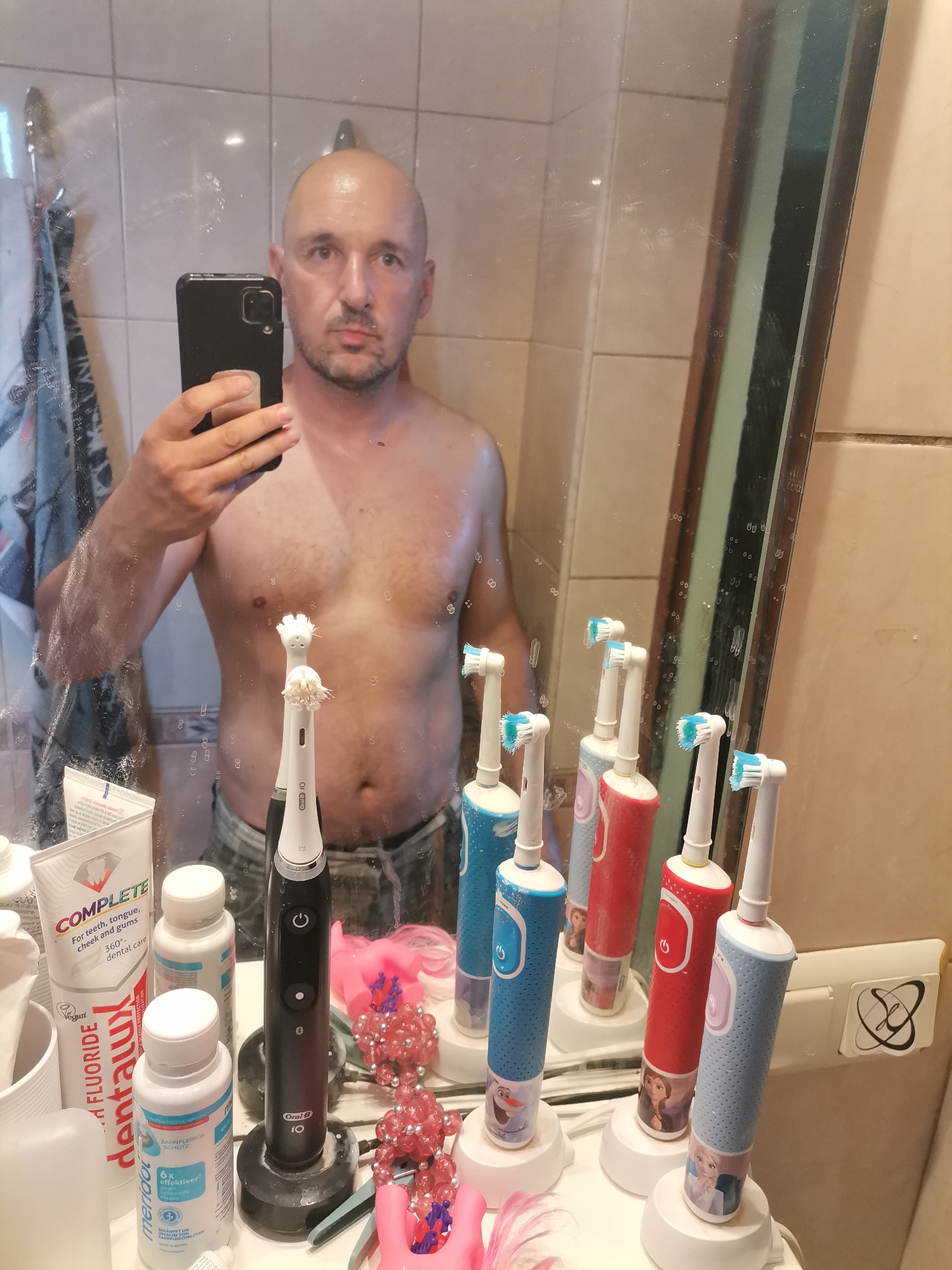 A partially undressed man is taking a selfie in a bathroom. He is bald, fair-skinned, and appears to be middle-aged. He is holding a smartphone in his right hand, presumably to take the photo. His torso is visible, revealing a bit of chest hair.
In front of him, on a bathroom counter, is an array of dental hygiene products. The products include toothpastes, mouthwash, and several electric toothbrushes. Some of the toothbrushes have characters on them, likely for children. A pink decoration made of beads and plastic is also visible.
The background reveals a tiled wall and a partially visible towel. The mirror in which the man is taking the selfie is splattered with water droplets. The scene appears to be a domestic bathroom setting.