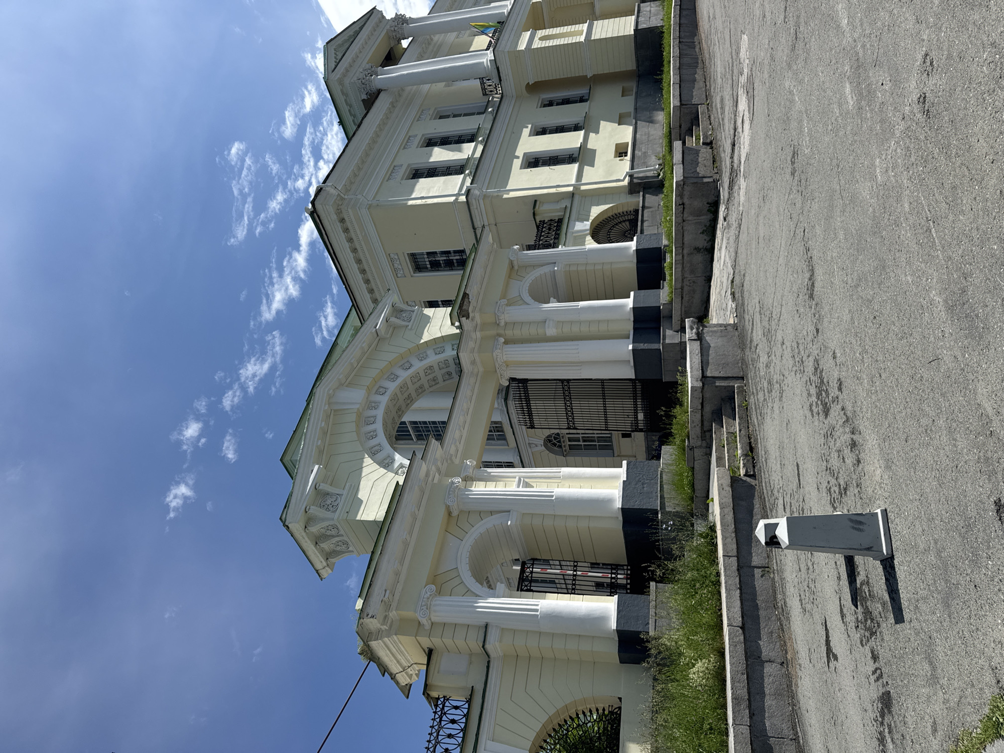 The image shows the exterior of a pale yellow neoclassical building with white columns and arched entrances, situated under a bright blue sky with scattered white clouds. The architecture features ornate detailing, a green roof, and large windows with black bars. In the foreground, a paved area with some grass and a light gray, truncated pyramidal bollard is visible. The building appears to be an official or historical site, possibly a museum or government building. Based on architectural style and common knowledge of Yekaterinburg landmarks, this is likely the "Old Believers' Church" or a similar structure in the city.
