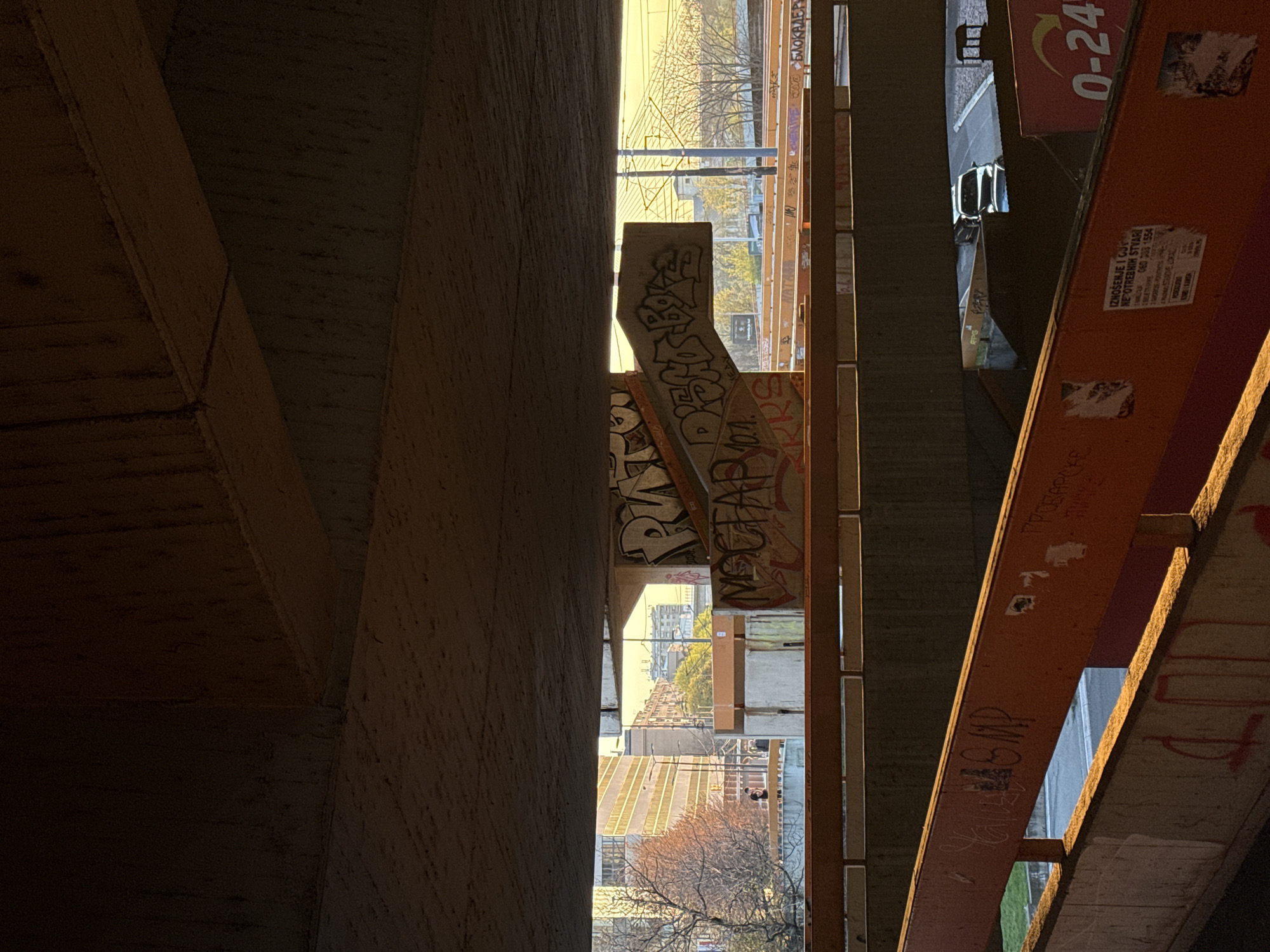 The image shows a view looking up through a gap between two concrete structures, likely an overpass or bridge in Belgrade, Serbia. Graffiti is visible on the concrete surfaces, including what appears to be the Serbian word "Blokade." One structure is reddish-orange and contains the marking "0-24" and a business contact advert. Through the gap, the cityscape is visible, including buildings, a street, and trees. A car is parked on a street above. The overall environment suggests a grimy, urban setting.