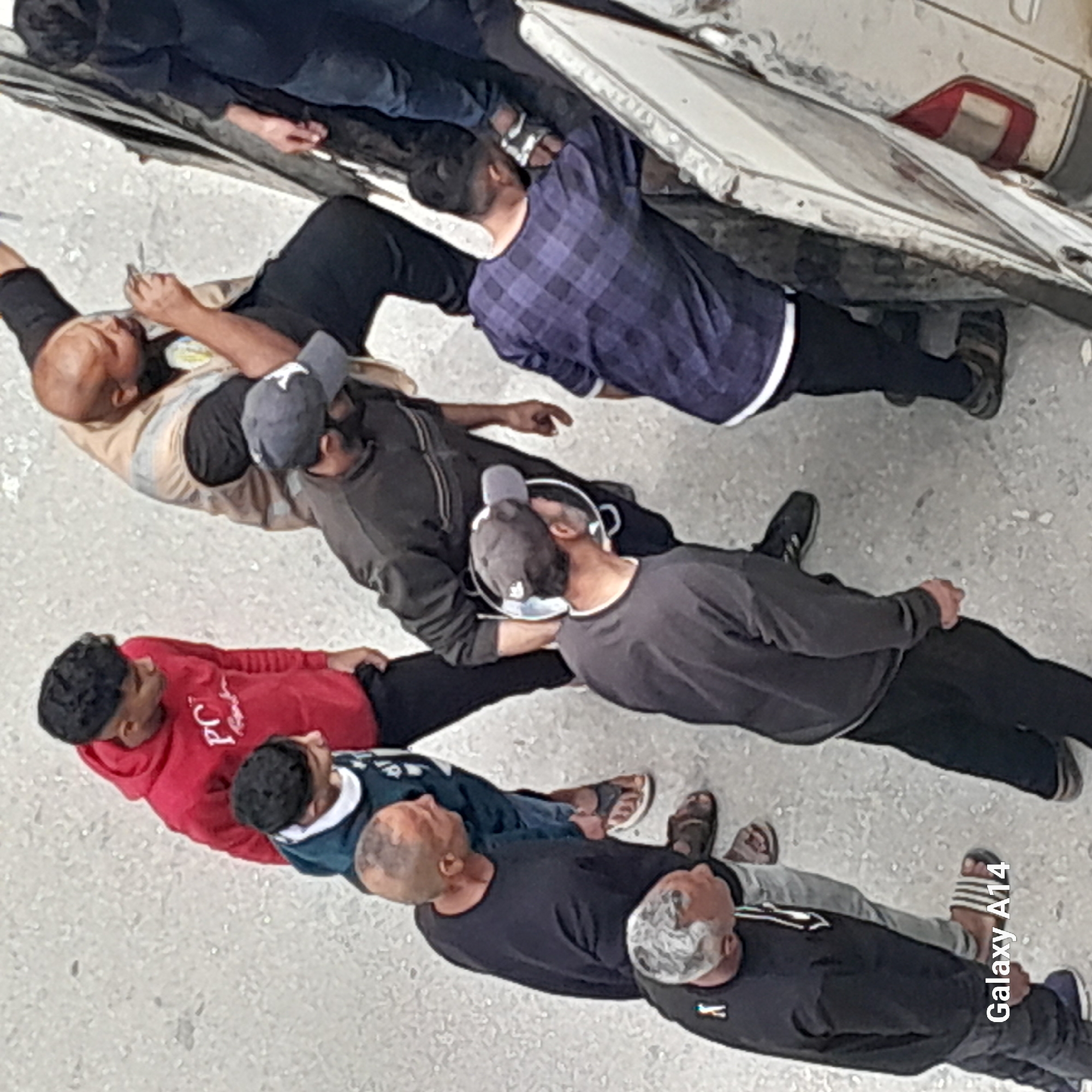The image shows a group of seven men seemingly lying on a grey-colored ground next to a white vehicle. The men are dressed in various casual clothing, including a red hoodie, a blue and white shirt, black shirts, and a plaid shirt. Some are wearing hats and sandals. The white vehicle is partially visible at the top of the frame. The location is specified as Unknown, Unknown.