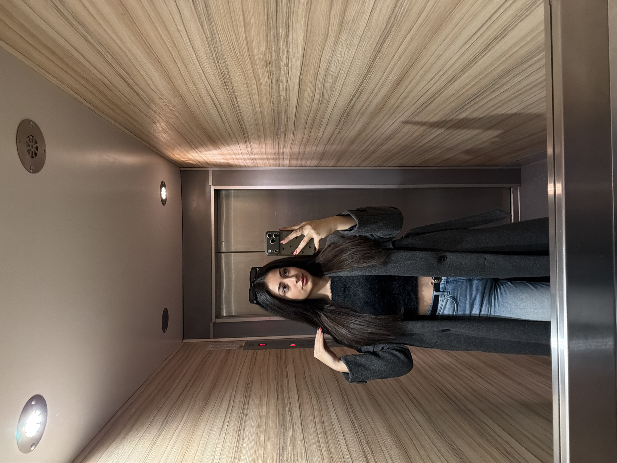 A woman stands in an elevator with wood-paneled walls, taking a selfie with her phone. She wears jeans, a black crop top, and a dark gray coat. Sunglasses sit atop her head. The elevator has a mirrored back wall and ceiling. The location is potentially in Sofia, Bulgaria, but this cannot be confirmed from the image alone.