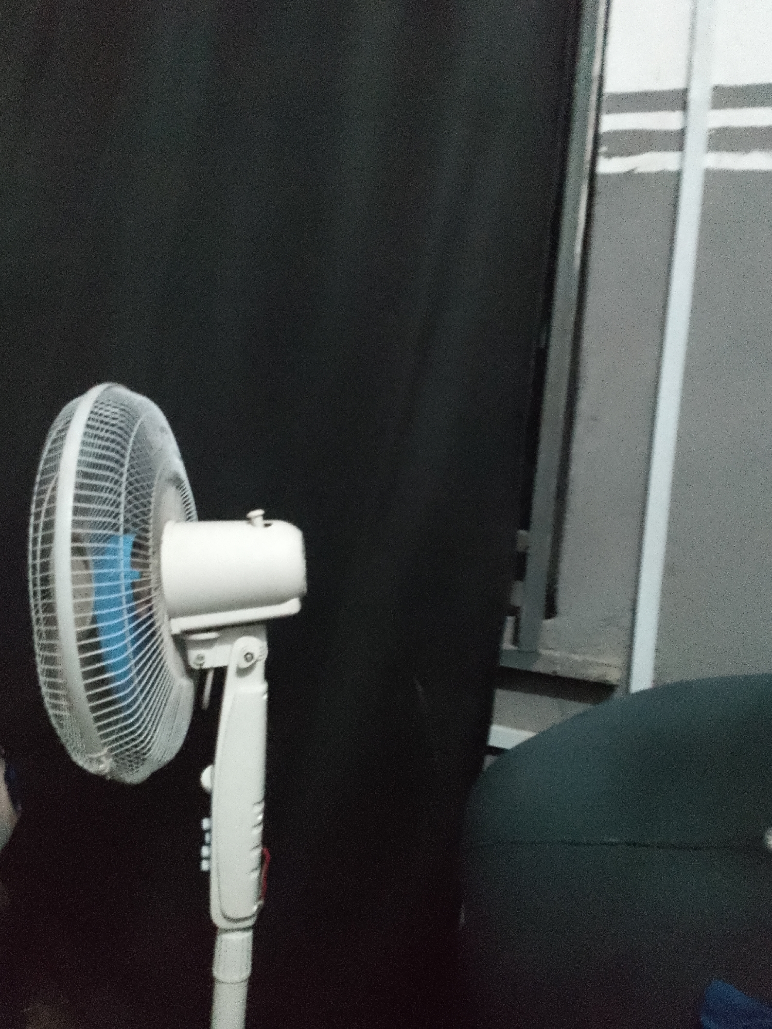 An indoor, medium-shot, eye-level, slightly blurry photograph shows a white oscillating fan positioned on a stand against a dark background. The fan has a circular cage with a white grill and a blue fan blade visible inside. The motor housing is also white, and a white stand supports the fan, extending downwards. To the right of the fan, a vertical gray surface with horizontal white stripes is visible, suggesting a wall or partition. In the foreground, on the right side of the image, a dark green, rounded object, possibly a piece of furniture, is partially visible. The lighting appears dim.