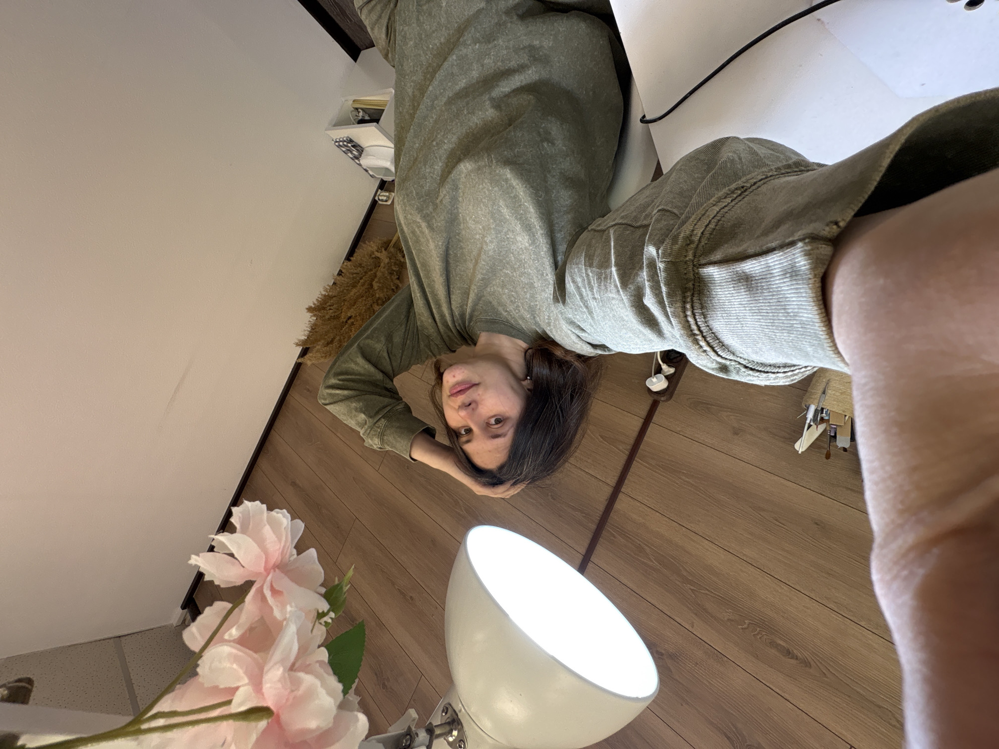 A woman with dark hair is lying on a wooden floor, wearing a green sweatshirt. Her head is resting on her hand. A white lamp is positioned near her. A bouquet of pink flowers is in the lower left corner of the image. In the background, there's a white wall and a shelf. Based on the available information, it's impossible to confirm whether the scene is in Chaikovsky, Russia.