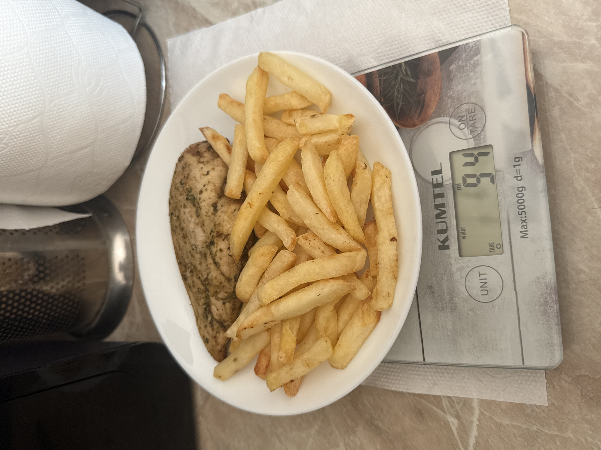 A white plate filled with french fries and a piece of seasoned chicken breast sits on a digital kitchen scale. The scale displays a weight of "94 ml". The scale is placed on a countertop next to a roll of paper towels and a dark appliance. The scene appears to be indoors, likely in a kitchen.