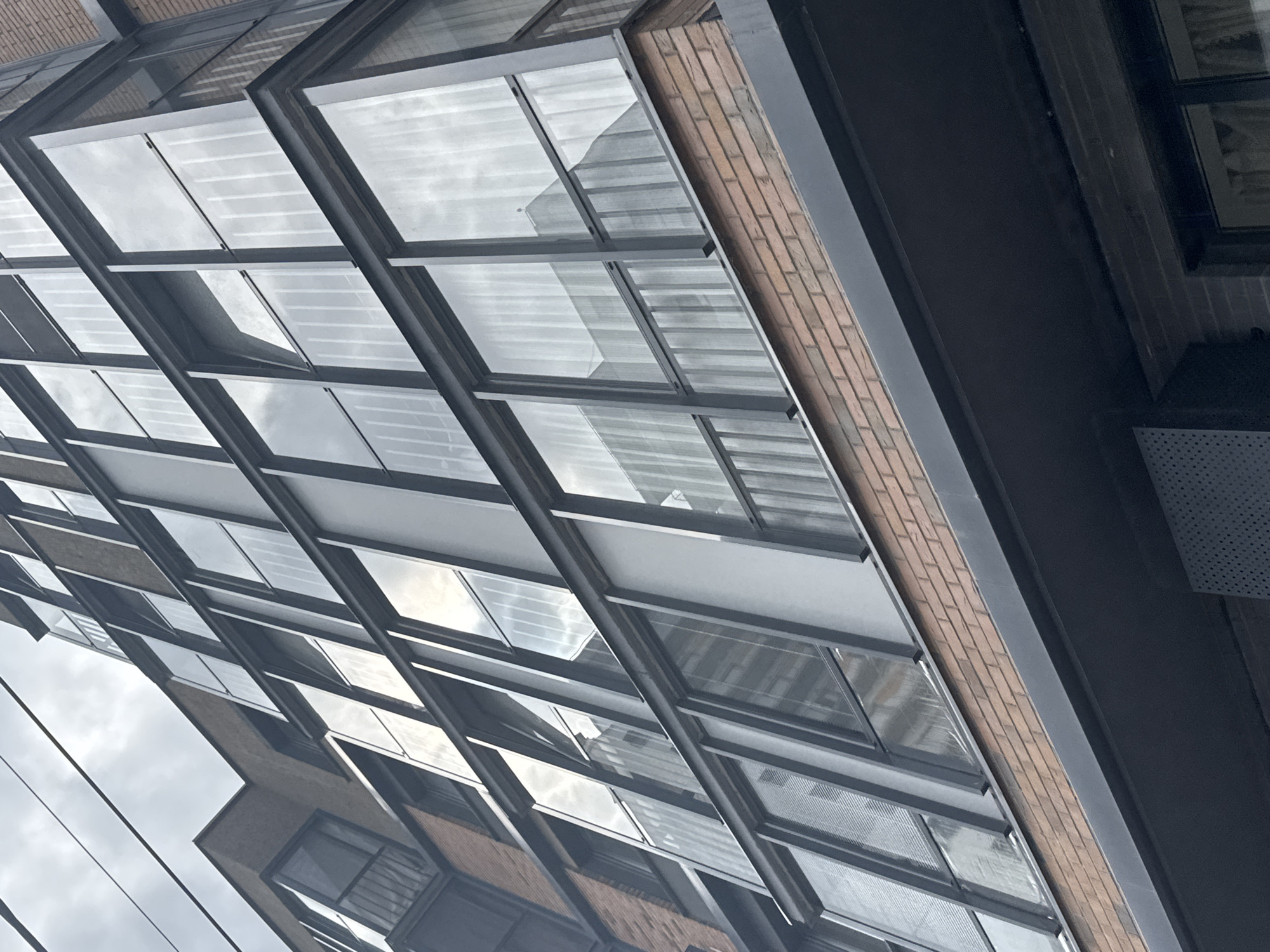 The image shows an angled view of a modern building exterior, characterized by a repetitive pattern of windows with dark metal frames. The windows appear to reflect a cloudy sky. A section of the building's facade is constructed of brick. Based on the visual architectural style, it might be located in the city of Perm, Russia.
