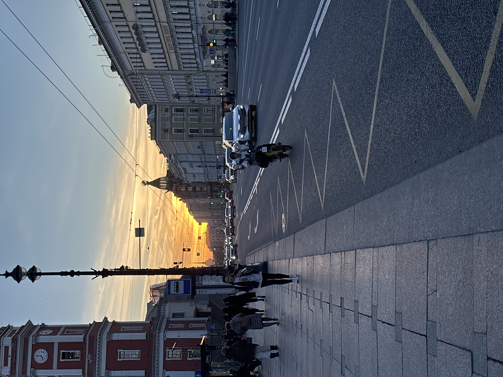 The image depicts a street scene in Saint Petersburg, Russia, during what appears to be sunset. The sky is a mix of orange and blue hues. On the left, there's a red brick building with a clock tower. A street lamp post stands in front of it. People are walking on the sidewalk. A street with multiple lanes runs to the right. Cars, a motorcycle, and other vehicles are on the street. The road markings are visible. Buildings line the street in the background. In the distance, the sunset creates a bright glow between the buildings.