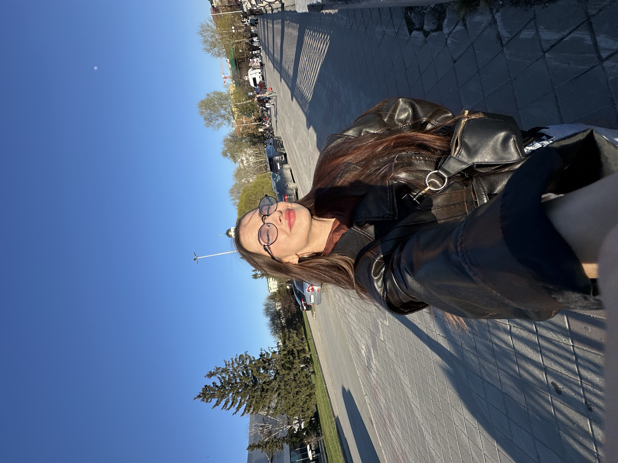 A selfie photograph captures a young woman outdoors on a clear, sunny day in what appears to be an urban or park-like setting in Yekaterinburg, Russia. The image is taken from a slightly low angle, showcasing a vast, bright blue sky where a faint moon is visible. Long, distinct shadows stretch across the grey block-paved ground, suggesting it is either morning or late afternoon.
The main subject is a young woman, shown from the chest up, with long, dark brown hair, fair skin, and round eyeglasses. She is wearing a dark brown or black leather jacket over a brown top. A small black shoulder bag with a silver ring and a visible zipper hangs from her shoulder. Her lips are slightly parted, and her expression is neutral to serene.
The background reveals a wide pedestrian area or street lined with a mix of deciduous trees (some bare, some with green leaves) and a large evergreen tree. Several parked cars and distant pedestrians suggest a lively public space. Various buildings are visible, including one with a distinct golden dome, possibly a church. A red construction crane can be seen far in the distance. No discernible text is visible in the image.