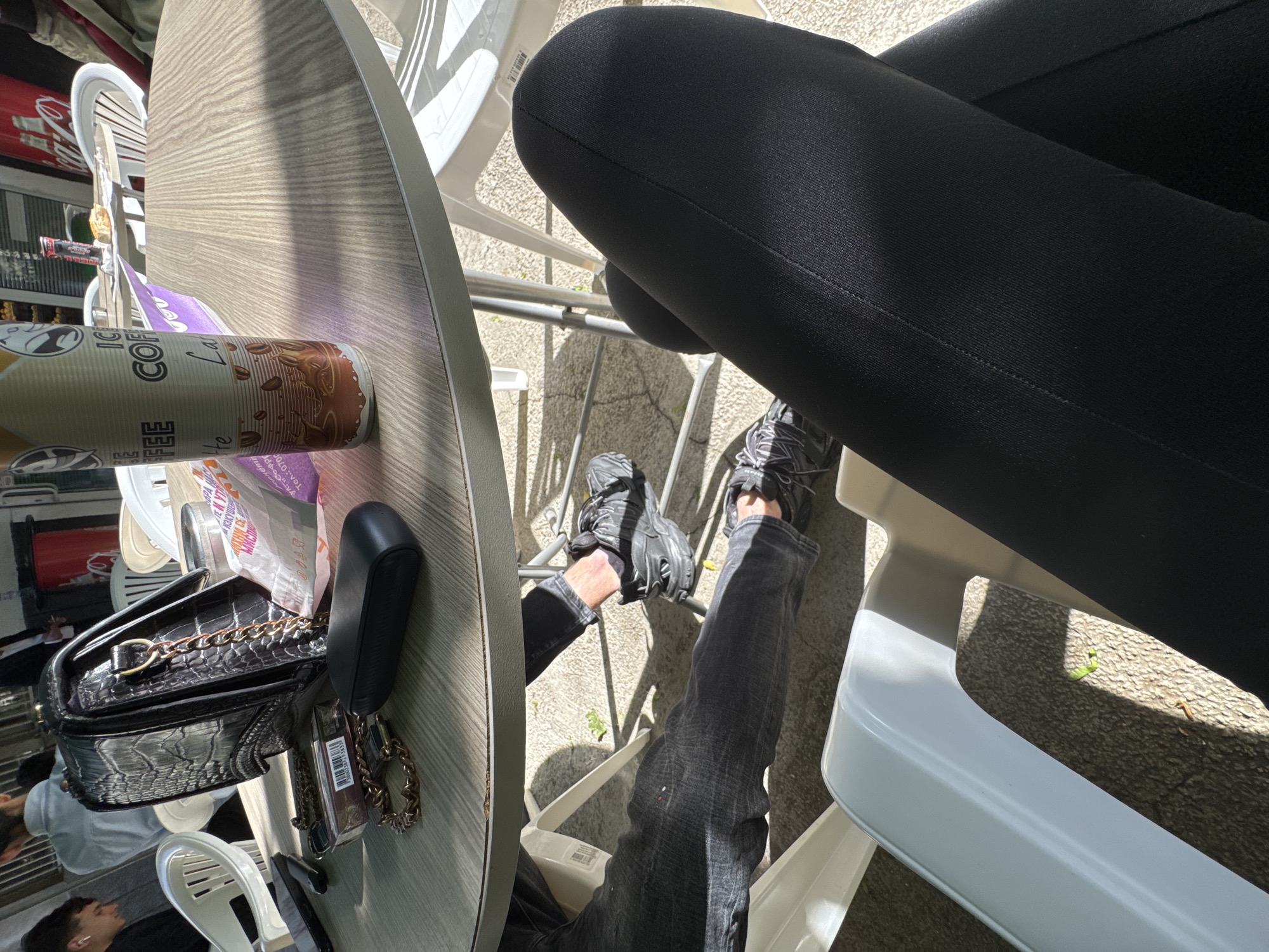A high-angle shot shows a person seated at a table in what appears to be an outdoor cafe setting. The person is wearing black leggings and gray denim pants, and a pair of black sneakers. On the table, there is a disposable coffee cup, an open packet of napkins, a black purse with a chain strap, and a black electronic device. The table has a faux wood-grain surface, and the person is seated in a white plastic chair. The location is likely in Sofia, Bulgaria, based on the Cyrillic text on the coffee cup. The image captures a candid moment, perhaps during a break or casual meeting. The scene suggests a relaxed and casual atmosphere. The composition focuses on the objects and the person's legs, providing a partial view of the environment.
