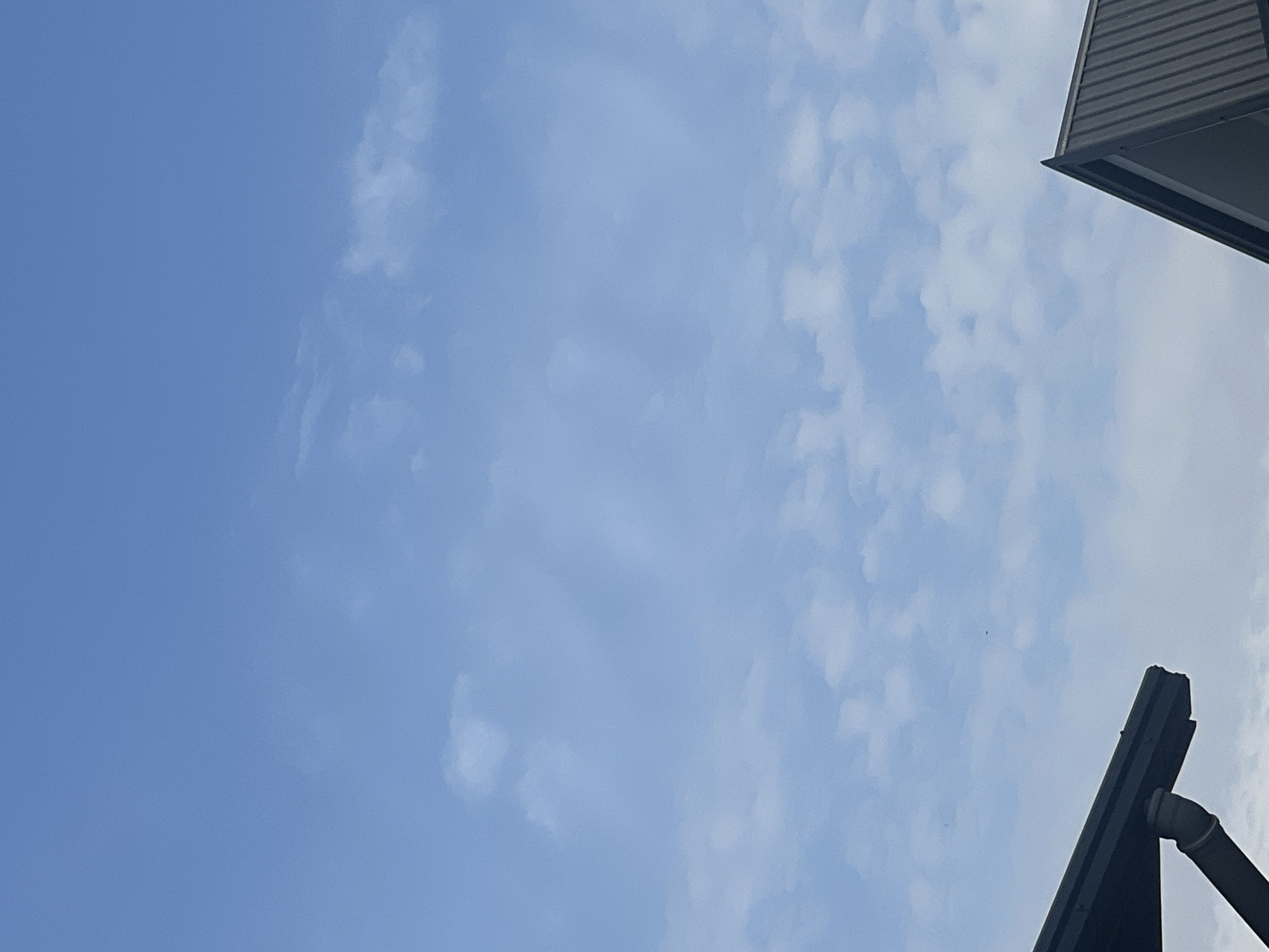 A close-up shot shows a clear blue sky with scattered white clouds. In the foreground, the dark, sloped roofs of two modern buildings are visible. One roof, on the left, is a dark overhang with a downspout attached. The other, on the right, is a pointed, dark gray roof with vertical seams. The image appears to be taken outdoors during daylight.