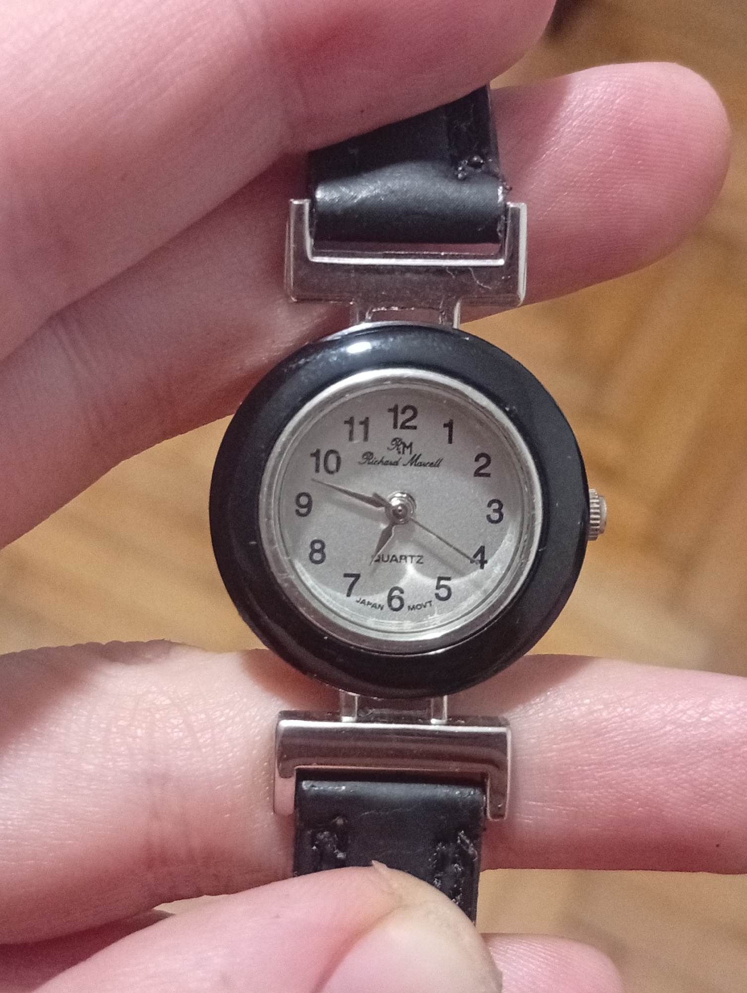 A wristwatch with a black bezel and a silver-colored face is held between two fingers. The watch face has numerals 1 through 12, and the brand name "Richard Maxwell" is printed below the 12. Below the center, the word "Quartz" is printed. The watch has a black leather strap attached with silver-colored metal hardware. The watch hands are positioned at approximately 10:21. "Japan Movt" is printed at the bottom of the dial below the six. The background is slightly blurred. There is no indication of the city of Buenos Aires, Argentina, or any recognizable location.