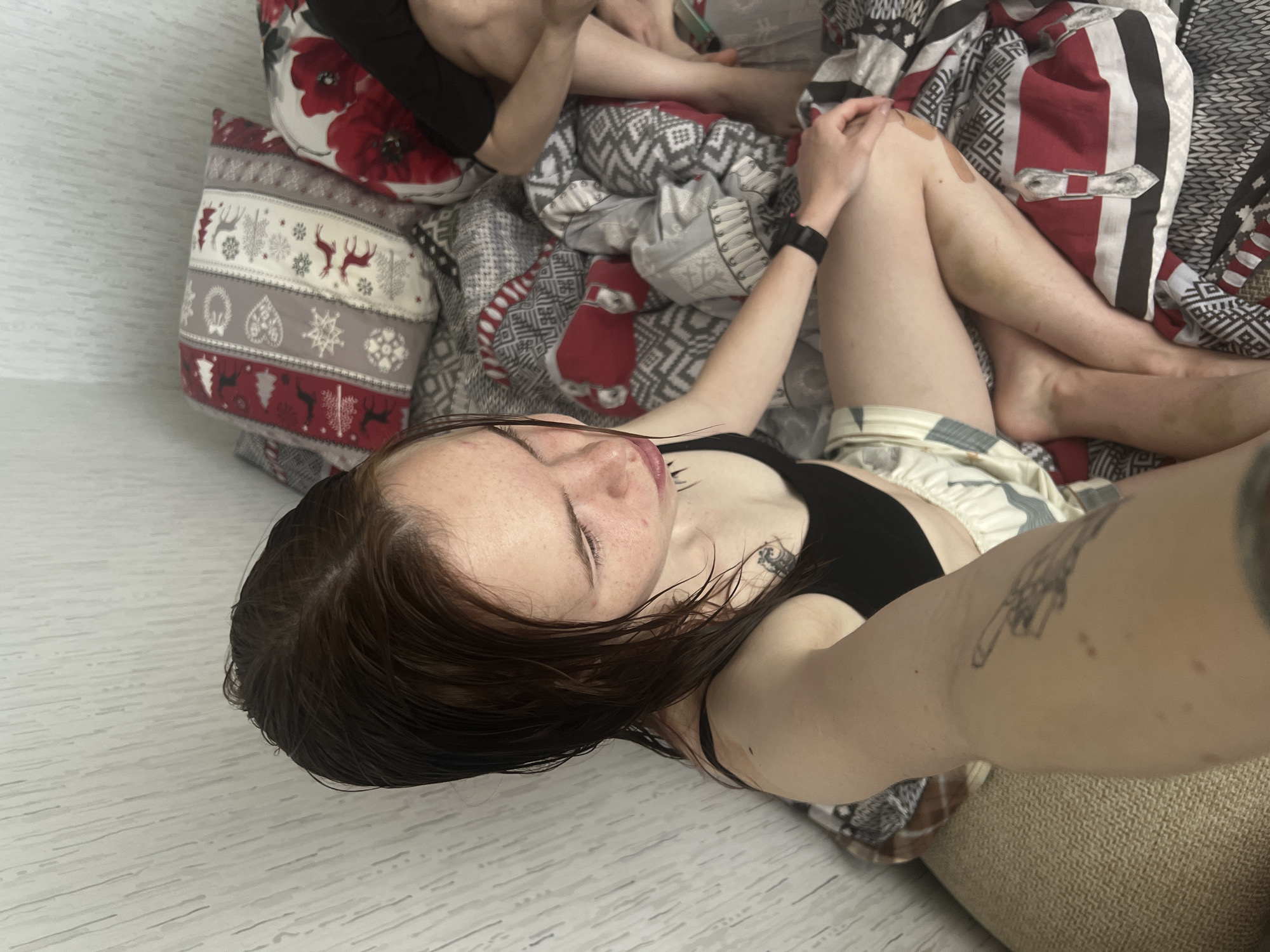 A selfie depicts a person with brown hair, wearing a black sports bra and patterned shorts, lying on a bed with patterned bedding and pillows. The person has tattoos on their arms. In the background, another person's legs are visible. The location is possibly Perm, Russia, based on the origin of the image.