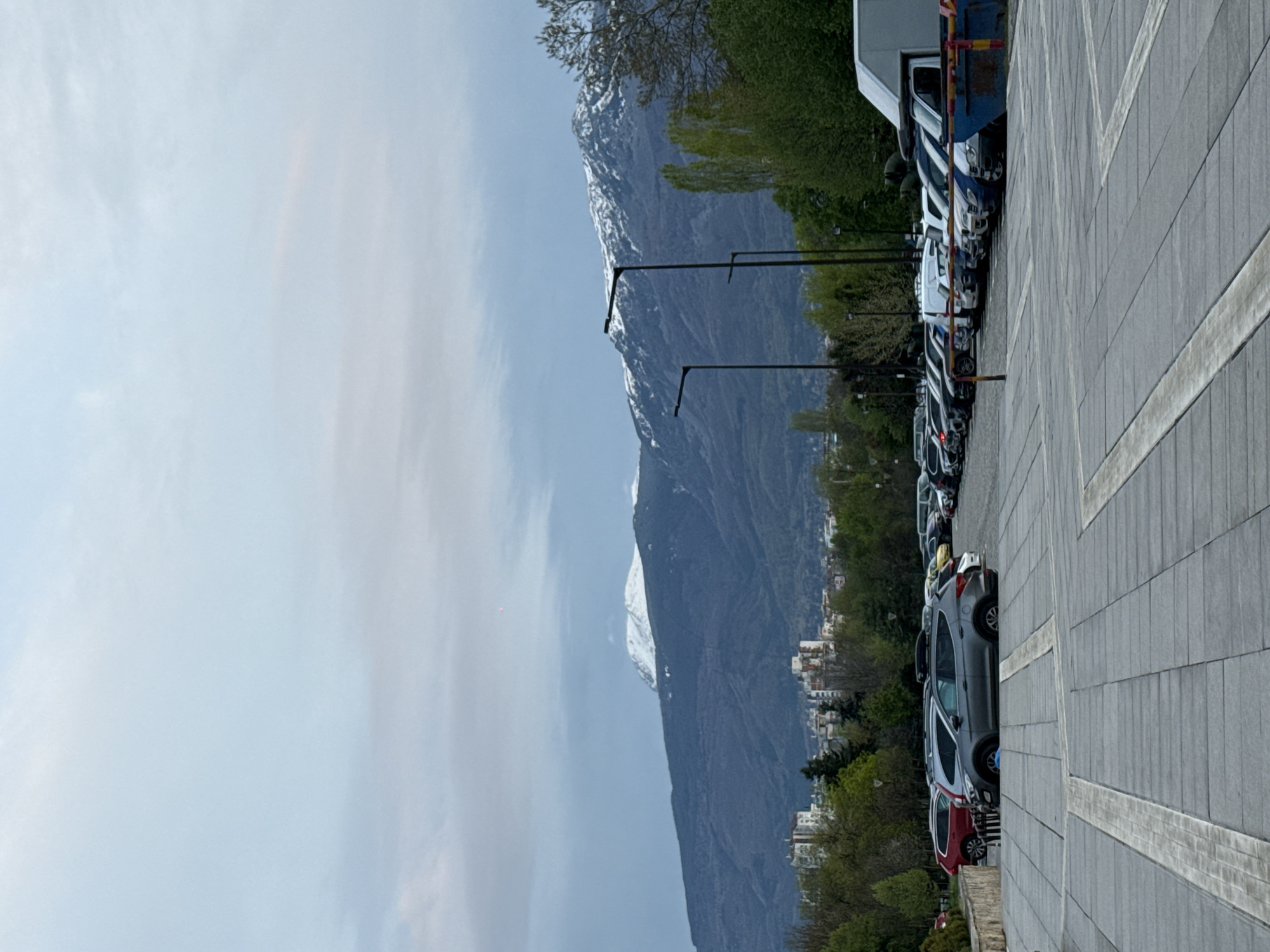 The image captures an outdoor scene dominated by a majestic mountain range in the background, set against a foreground featuring a parking lot and paved area. The photograph is rotated 90 degrees clockwise.
In the foreground, a wide, grey paved surface, possibly a sidewalk or part of a plaza, occupies the right side and bottom of the frame. Distinct lighter-colored stripes run across this paving. Adjacent to this, a row of parked vehicles stretches across the middle ground. These include a white van or truck with a blue and yellow striped back, a grey SUV, a red car, and several other vehicles of varying colors and models.
Beyond the vehicles and further towards the left (as the image is oriented), a line of lush green trees partially obscures the base of a large, imposing mountain range. The mountains are dark and rugged, with significant snow patches visible on their higher peaks, suggesting a high altitude or a temperate climate with recent snowfall. Scattered between the parked cars and the mountains are several tall, dark streetlights or utility poles. Faintly visible at the foot of the mountains, nestled among the trees, are distant buildings and residential structures.
The sky is overcast and a soft blue-grey, indicating diffused daylight, possibly late morning or early afternoon, with no harsh shadows. No people are present, and no specific activity or event is taking place beyond the static parking of vehicles. No text is visible in the image. The scene blends urban elements with striking natural beauty, set in Sofia, Bulgaria.