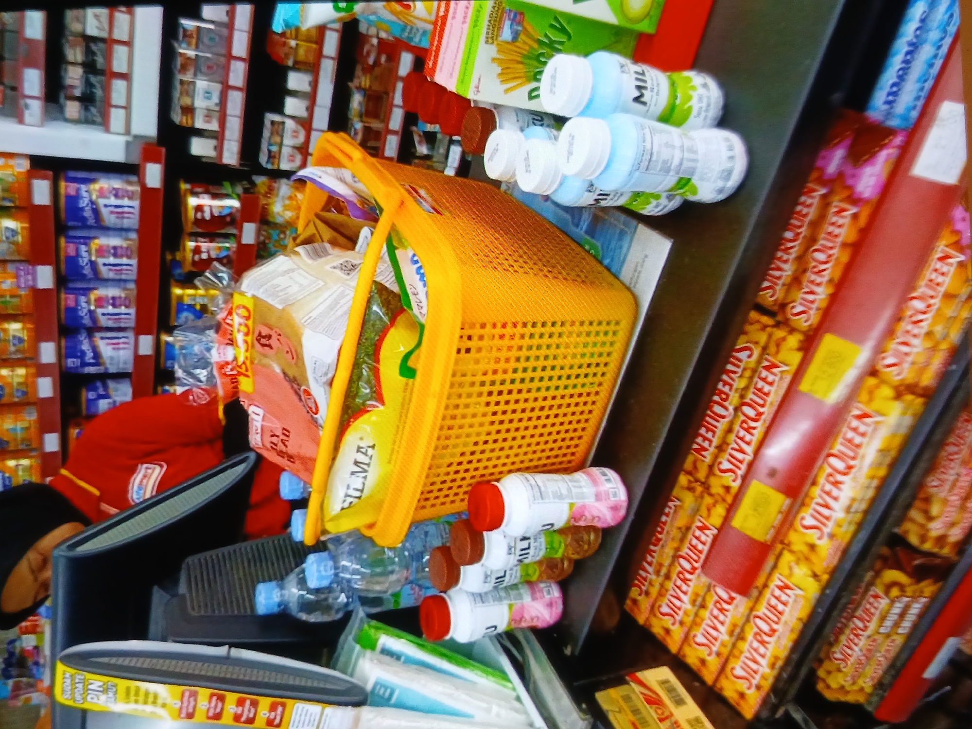 In a brightly lit convenience store in Setia Bakti, Indonesia, a female cashier wearing a red uniform with a yellow collar and a black hijab is partially visible behind a computer monitor, attending to a transaction. In the foreground, a vibrant orange plastic basket is filled with various grocery items, including packets of food and what appear to be bottles of beverages. To the left of the basket, several small bottles with red and brown caps are neatly arranged on the counter, along with more bottled drinks. The counter itself displays an assortment of packaged snacks and candy bars, prominently featuring the "SilverQueen" brand. The shelves in the background are stocked with a variety of packaged goods, including bottles, boxes, and tins of food items. The overall scene depicts a typical retail environment within the store.