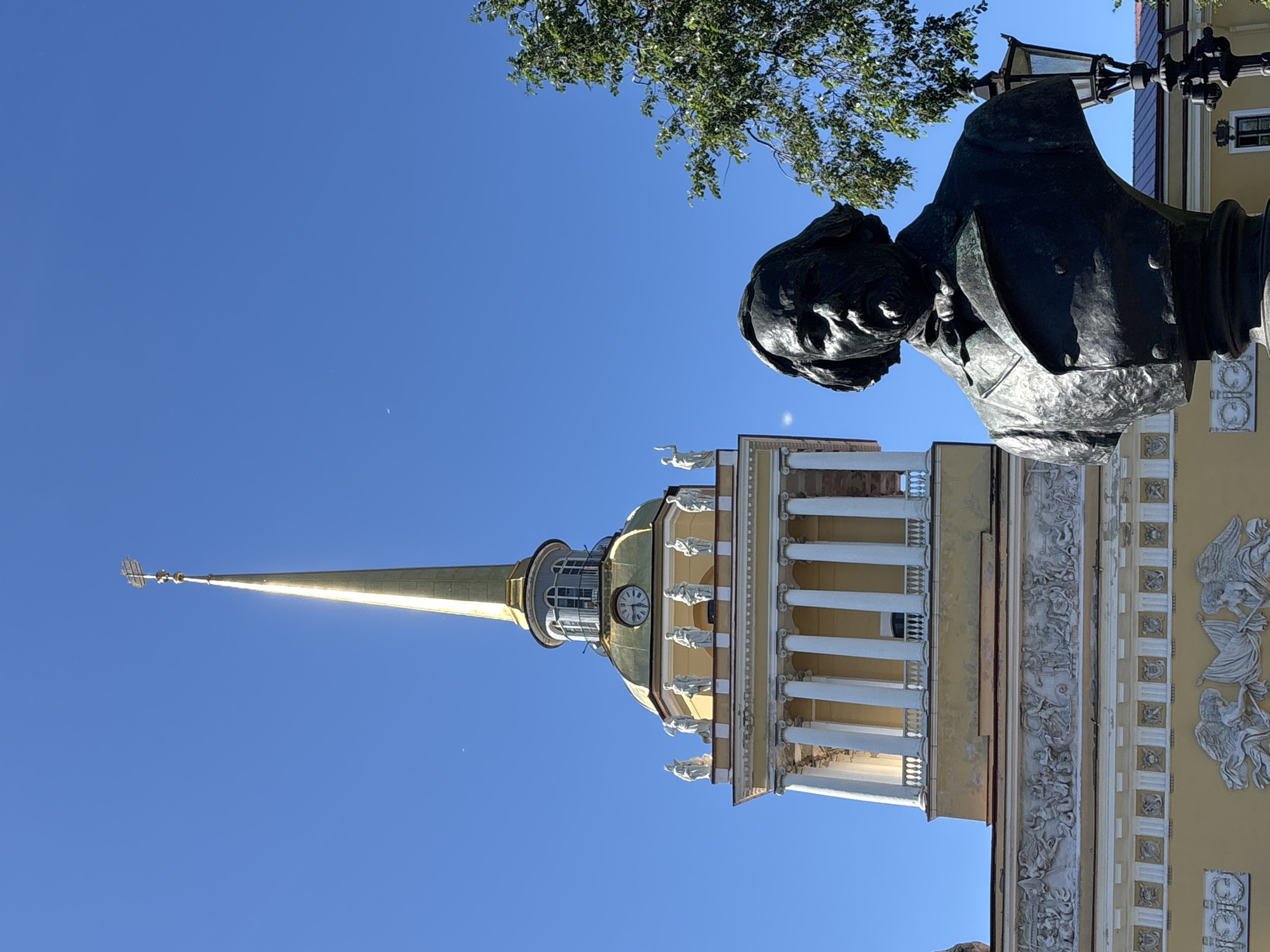 This image captures a bust of a man in the foreground, facing right, with his head and shoulders visible. He has a beard, a bow tie, and is wearing a double-breasted jacket. Behind the bust, a tall, yellow building with white columns and statues on top is visible, topped by a golden spire. The building is likely the Admiralty Building in Saint Petersburg, Russia, due to its distinctive golden spire and architectural style. The sky is a clear, bright blue, with a few faint specks that might be dust or distant clouds. A tree branch with green leaves extends into the frame from the right side of the image. The overall scene appears to be a sunny day in a public space.