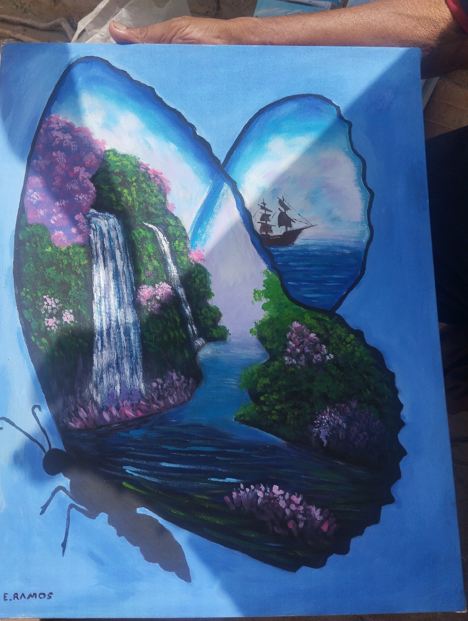 The image shows a painting of a butterfly with scenes inside its wings. The left wing depicts a waterfall cascading down a verdant, tree-covered hillside with pink flowering plants. The right wing contains an image of a sailing ship on the ocean, with similar greenery and flowering plants along the shore. A river connects the two scenes. The butterfly's body and antennae are shadowed, and the background is a solid light blue. The signature "E.RAMOS" is visible in the lower left corner of the painting. It is not possible to determine if this image is from the city of El Piache, Venezuela or the location from the photograph.