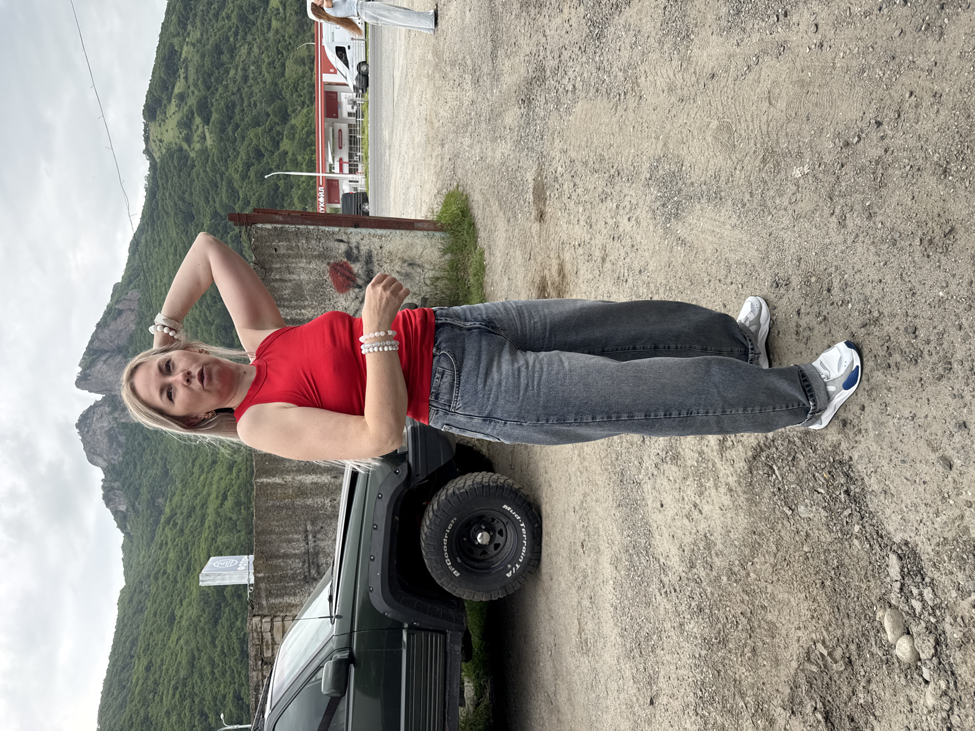 A woman with blonde hair and fair skin is posing next to a dark green SUV, with a mountainous landscape in the background. She is wearing a red sleeveless top, dark wash jeans, and white sneakers. She is adorned with several beaded bracelets on her left wrist. Her pose is suggestive, with her right arm bent and hand on her head. The SUV's tire is visible, and it appears to be a mud-terrain tire. In the background, there is a building with red and white accents, possibly a gas station, and beyond that, a lush green mountain under a cloudy sky. A second person, mostly out of frame, is standing near the building. The ground is a mix of dirt and gravel. The image seems to be taken during the day, with overcast weather. The Russian text "ЛУКОЙЛ" (LUKOIL) is visible on the building. The scene is set in Karachayevsk, Russia.