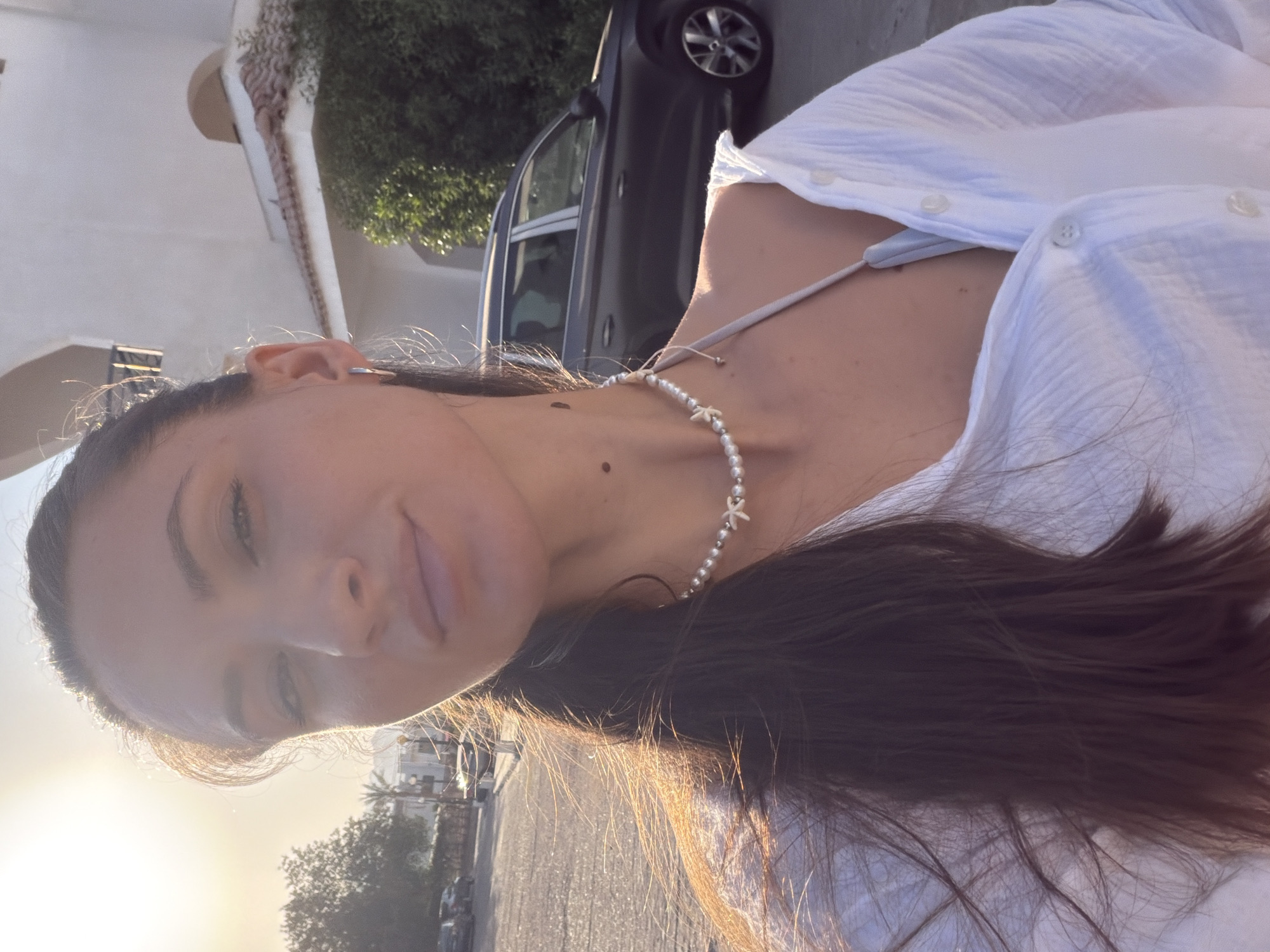 A woman stands outdoors, facing the camera in a selfie. She has long, dark hair that is windswept around her face and shoulders. She is wearing a white button-up shirt, partially unbuttoned, revealing a light-colored strap of undergarment underneath. A pearl necklace with a small starfish pendant adorns her neck. The background shows a paved street with parked cars and a building with a tiled roof and archways. Lush green bushes are visible. The sun is low in the sky, casting a warm glow on the scene, suggesting it is either sunrise or sunset. This scene is consistent with the environment found in Sharm El Sheikh, Egypt, a popular tourist destination known for its coastal resorts and sunny climate.