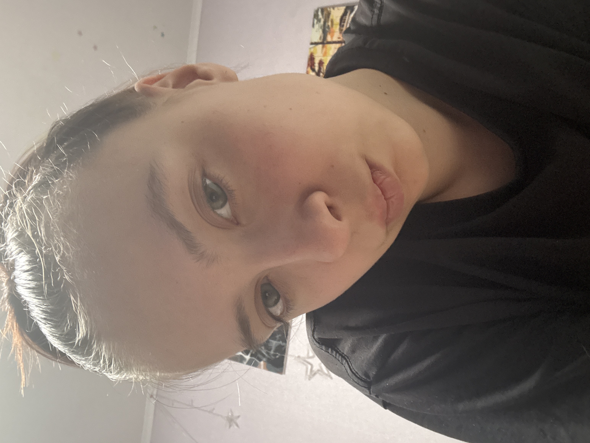 A close-up, angled shot of a person with fair skin, green eyes, and light brown hair pulled back from their face. The person is wearing a black shirt. The background includes a light-colored wall and some indistinct objects, possibly decorations. The location given is Прикамский, Russia, but this cannot be verified from the image.
