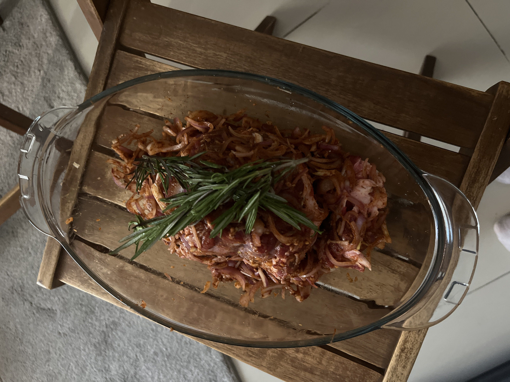 A close-up, high-angle shot shows a clear glass baking dish filled with raw marinated meat and sliced red onions, topped with a sprig of rosemary. The dish sits on a slatted wooden table, which is positioned on a grey carpet. The background is out of focus, but appears to be an indoor setting with a wall and flooring. There is no discernible information about the location in Dubai, United Arab Emirates.