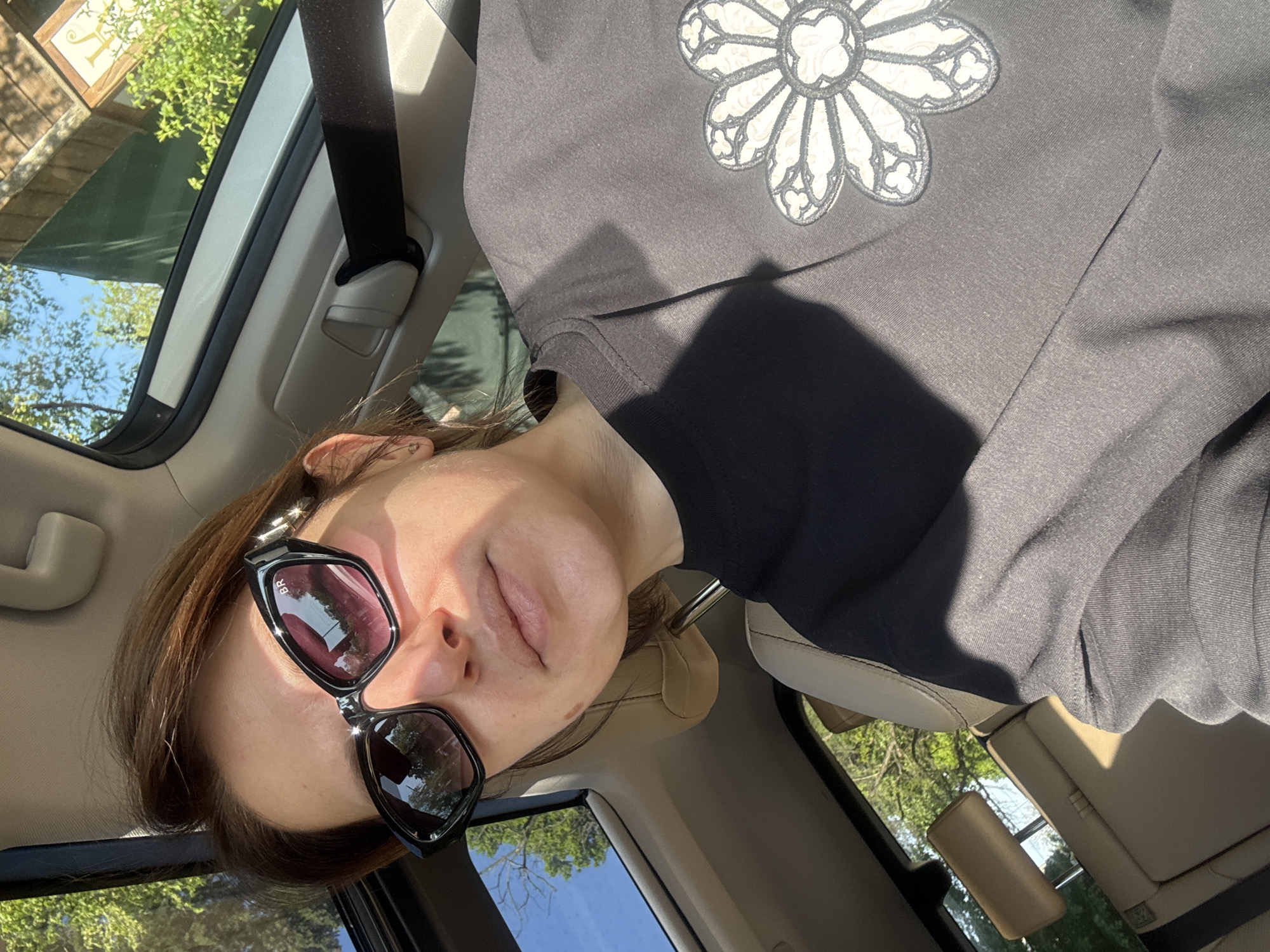 A woman wearing sunglasses and a black top with a floral design is seated in a car. The vehicle's interior includes a tan seat, a seatbelt, and a sunroof. The view through the sunroof shows trees and a blue sky. The location of the image cannot be determined to be Sofia, Bulgaria based on the available visual information.