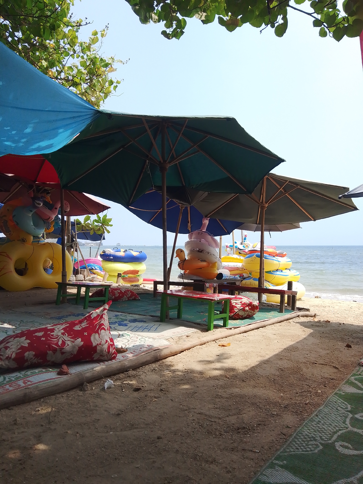 The image depicts a beachfront scene in Pejagan, Indonesia, on a sunny day. Several large, colorful umbrellas provide shade over a sandy area. Underneath the umbrellas are tables with green legs and red tops, and colorful inflatable rings are stacked on the beach. In the foreground, a red and white floral patterned cushion lies on a mat. The ocean is visible in the background, with a clear blue sky above. Trees with green foliage frame the top of the image. There are no people visible in the frame.