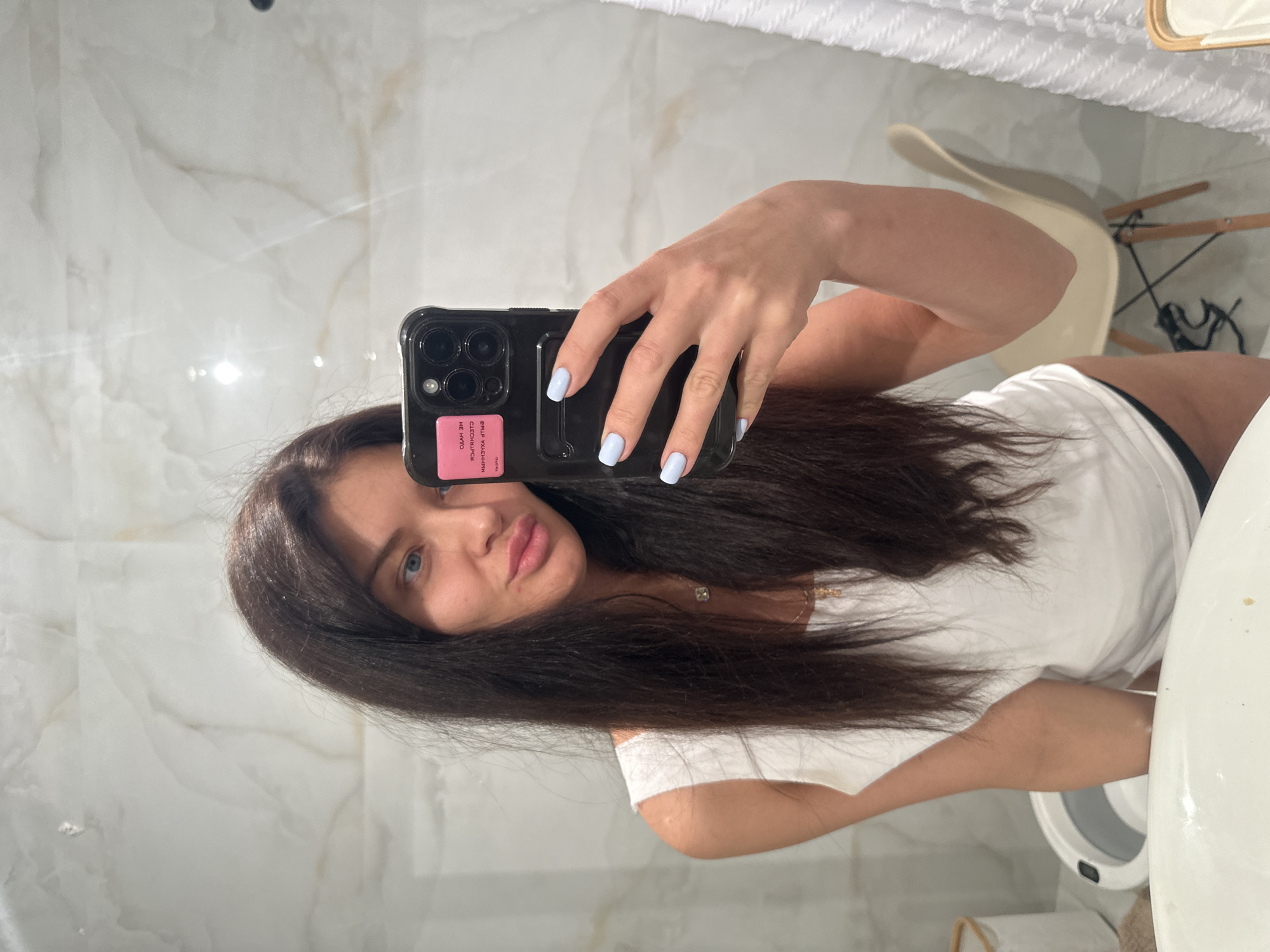 A brunette woman is taking a selfie in what appears to be a bathroom. She is holding a black smartphone with a pink sticker on the back. Her nails are painted light blue. She is wearing a white t-shirt. The bathroom has white marble-like walls and a white toilet is visible. A light-colored chair is in the background. The scene does not appear to be in the city of Khimki, Russia.