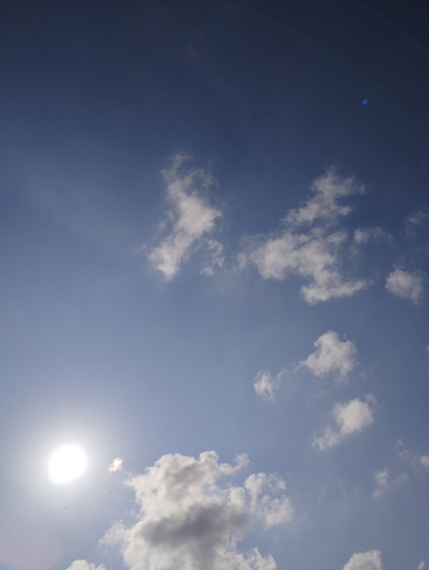 The image displays a clear and vibrant blue sky, predominantly filled with sunlight and scattered clouds. The sun, appearing as a brilliant white, overexposed orb, is positioned in the lower-left portion of the frame, casting visible rays of light into the atmosphere. This strong illumination suggests a bright daytime setting, likely mid-morning to late afternoon.
Various white, fluffy cumulus clouds are distributed across the sky. Some clouds appear intensely bright due to the direct sunlight, while others further from the sun, particularly in the lower-central and right areas, show subtle grey shading, indicating their three-dimensional form.
A distinct blue circular lens flare is noticeable in the upper-right corner of the image, a common artifact when photographing directly into the sun. A few very tiny, dark specks, possibly distant birds or dust on the camera sensor, are barely visible against the blue sky, particularly near the sun and in the upper-mid section. There are no specific location cues within the image itself that would identify Barcelona, nor is any text visible. The overall scene conveys a sense of serene and sunny weather.