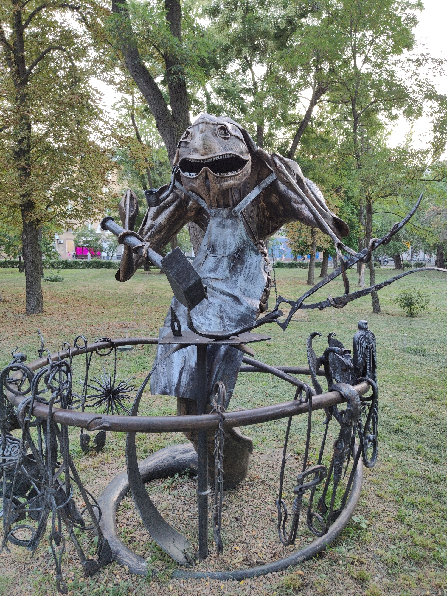 A metal sculpture of a creature resembling a reptilian blacksmith stands in a park in Donetsk, Ukraine. The creature, with its mouth open in a wide grin and large, round eyes, wears an apron and holds a hammer in its right hand. It is surrounded by a decorative metal fence adorned with various metalwork pieces, including what appears to be a spiked ball and chain-like elements. The sculpture is situated on a patch of ground covered with gravel and grass, with trees and a building visible in the background. The overall setting suggests an outdoor public space, possibly a park or a square.