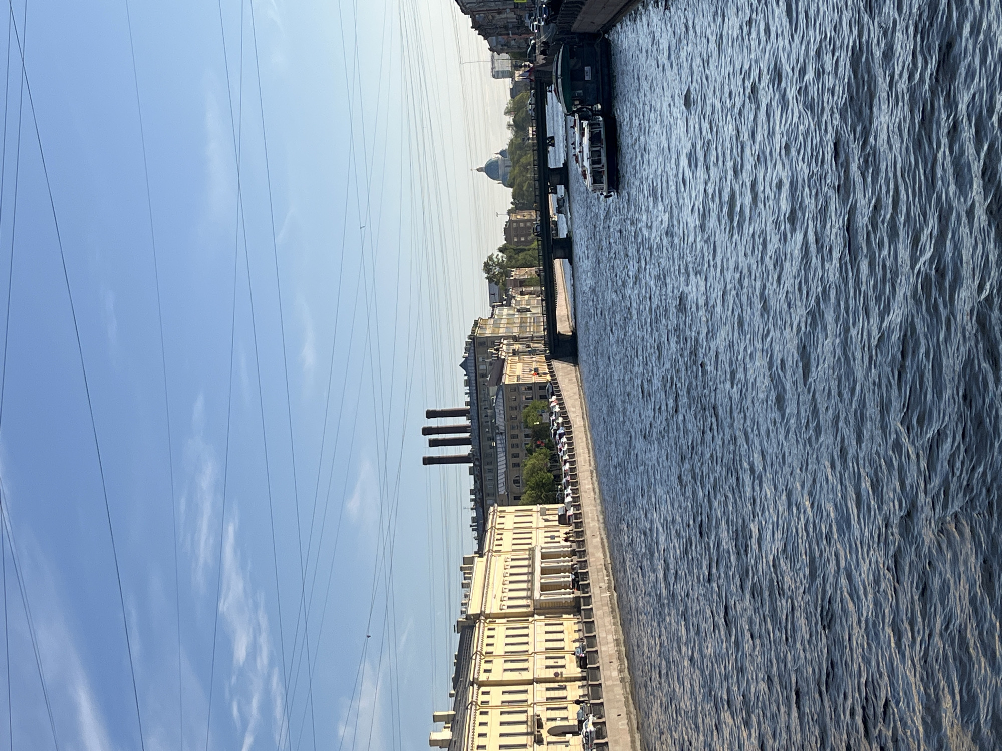 The image depicts a waterway in Saint Petersburg, Russia. On the left is a pale yellow building facade running alongside the canal with three tall industrial smokestacks further down the waterway. Numerous overhead power lines run across the sky. The water is choppy with visible ripples. A bridge spans the waterway further in the distance, beyond which a large dome-shaped building is visible. Several boats are docked along the far side of the canal. Cars are parked along the embankment on the same side as the smokestacks. The sky is blue with scattered wispy clouds.