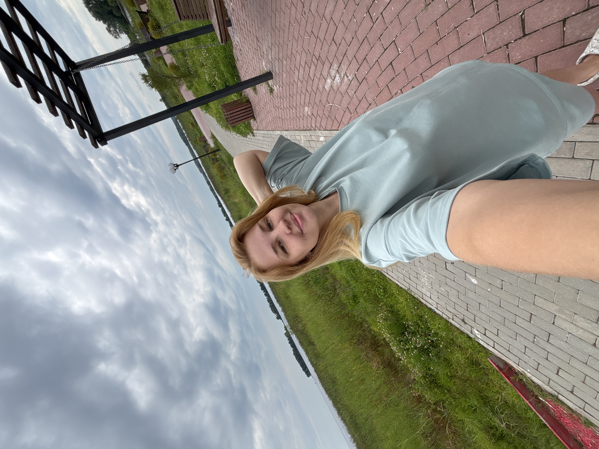 A woman with blonde hair takes a selfie outdoors in Breytovo, Russia. She is wearing a light blue t-shirt and stands on a paved walkway. To her left is a grassy area bordering a body of water. To her right, there is a wooden pergola with a swing, benches, and a trash can. The sky is cloudy.
