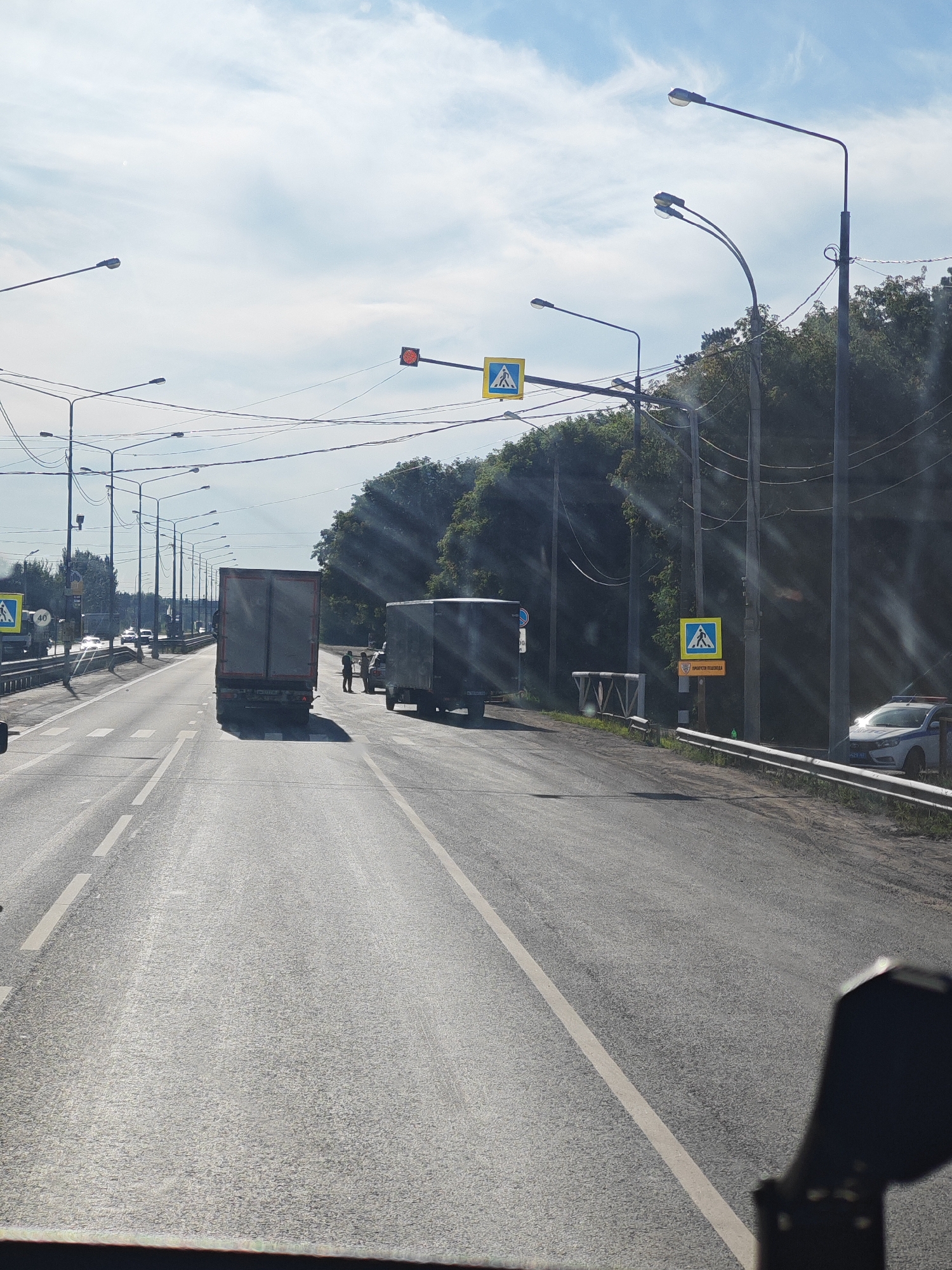 The image depicts a roadside scene in Verkhnee Sancheleevo, Russia, on a sunny day with a partially cloudy sky. The road is a multi-lane asphalt highway. In the foreground, the perspective is from inside a vehicle, as indicated by a dark object on the lower right edge, possibly a side mirror.
The primary focus is on the road and the surrounding environment. Two large trucks are visible on the road. The truck in the center is a grey semi-trailer truck facing away from the viewer. To its right, another grey box truck is parked. Several people, appearing to be two or three individuals, are standing near the parked box truck.
Traffic lights are present, with one showing a red signal. Several streetlights line the road, supported by poles and interconnected by wires. Pedestrian crossing signs with the international symbol of a person walking are also visible. One such sign is prominent on the right side of the road, accompanied by another sign with Cyrillic text. A white police car with flashing lights is parked on the right shoulder of the road, partially obscured by a metal guardrail.
The background consists of dense green trees and foliage, suggesting a rural or semi-urban setting. The road markings are clear, with white lines dividing the lanes. The overall impression is a typical daytime scene on a highway in Russia.