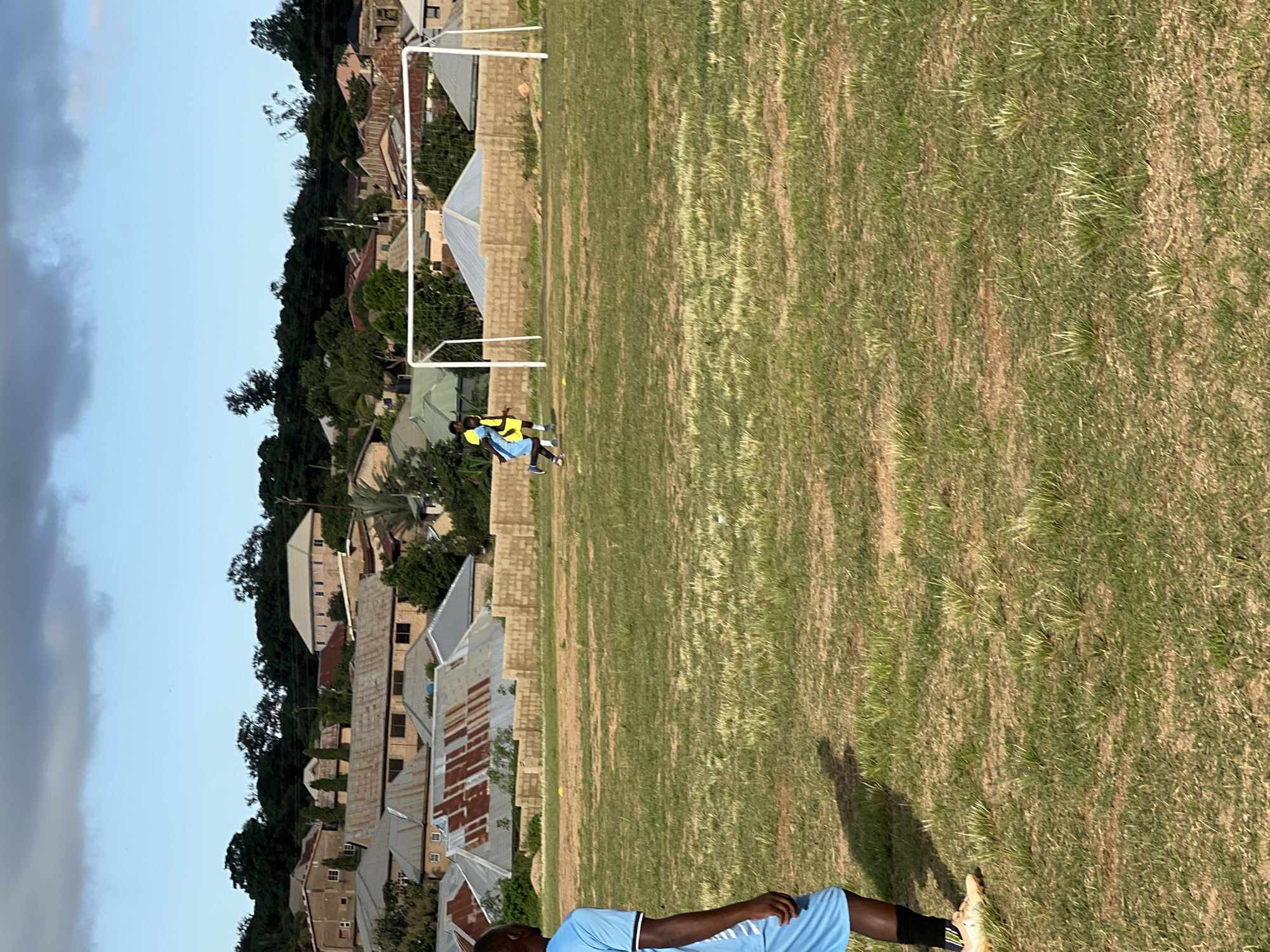 An outdoor, low-angle, eye-level, medium shot captures a grassy, uneven football field in Jos, Nigeria, on a clear day with a partly cloudy sky. In the foreground, the left side of a person wearing a light blue t-shirt and light blue shorts with black socks and beige cleats is visible, with their back to the camera. The person is standing on the field, casting a long shadow to their left. In the middle ground, two individuals are positioned near a white football goalpost. One individual, wearing a bright yellow vest over a blue shirt and shorts, is running towards the goal. The other individual, wearing a blue shirt and shorts, is standing behind the goal. A low, tan brick wall with scattered vegetation runs horizontally across the middle ground. In the background, a densely populated residential area is visible, with buildings featuring corrugated iron roofs and varying architectural styles. Trees and vegetation are interspersed among the houses, and a sloping, tree-covered hillside forms the furthest background.