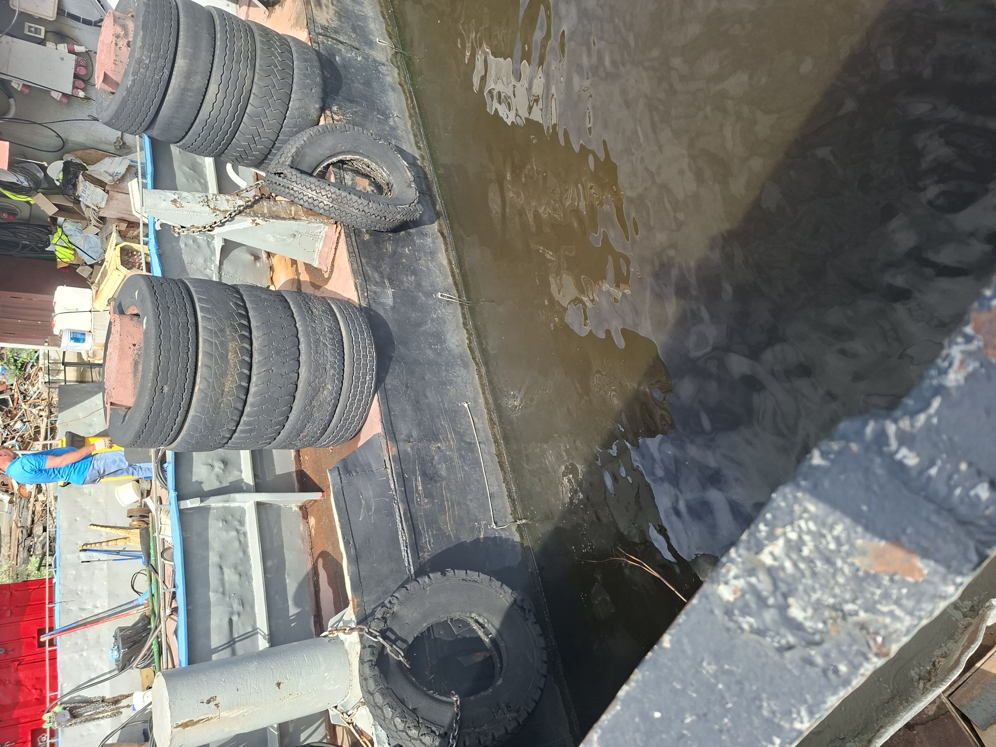 The image shows a view from a boat or barge docked on the Kazanka River in Kazan, Russia. In the background, a person in a blue shirt and jeans is visible, standing near some debris. The foreground of the image features several stacked tires and one tire hanging off the side of the vessel, used as fenders. The water is murky brown, reflecting the sky and the vessel. The overall impression is one of industrial activity or storage near the river.