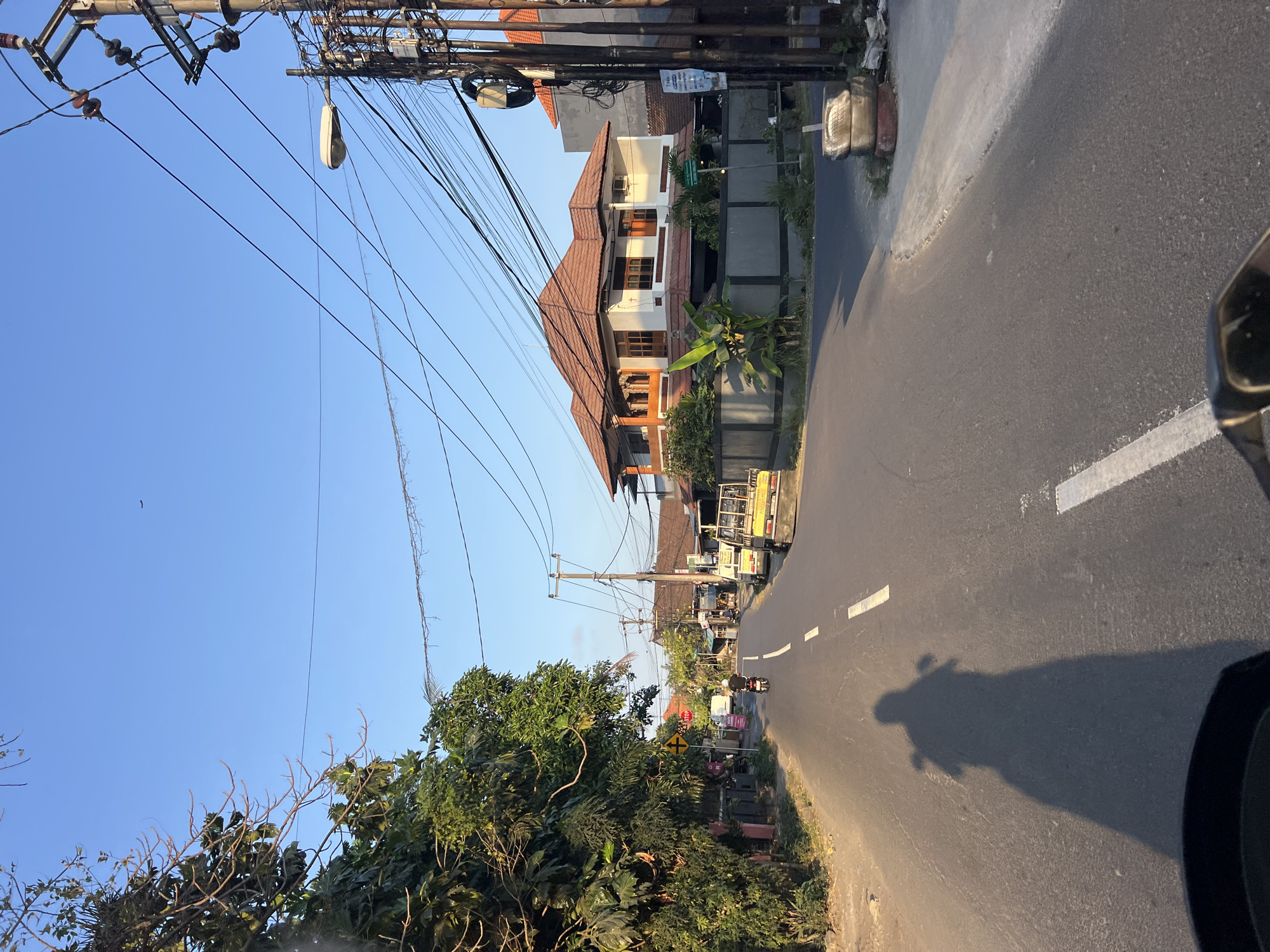 A motorcycle shadow is cast on an asphalt road in Denpasar, Indonesia. The motorcycle is being ridden by a person wearing a helmet. The road is lined with trees on the left and buildings and utility poles on the right. The sky is clear and blue.