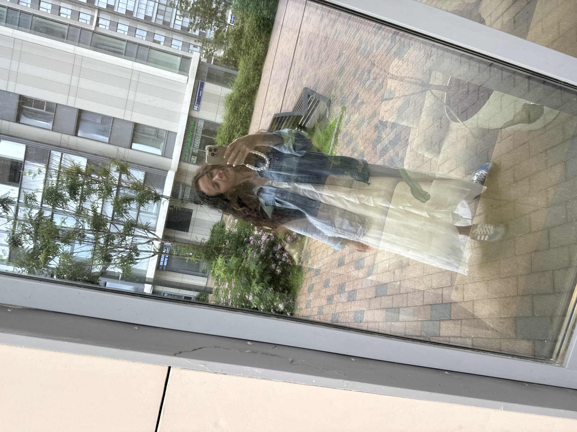 A woman is taking a mirror selfie in front of a modern apartment building in Moscow, Russia. She is wearing a light-colored maxi dress, a denim jacket, and sneakers. In the reflection, we see a tree with pink flowers, a brick-paved courtyard, and a bench. The building behind her has a facade with large windows and geometric elements. The image is taken during the day, with ample natural light.