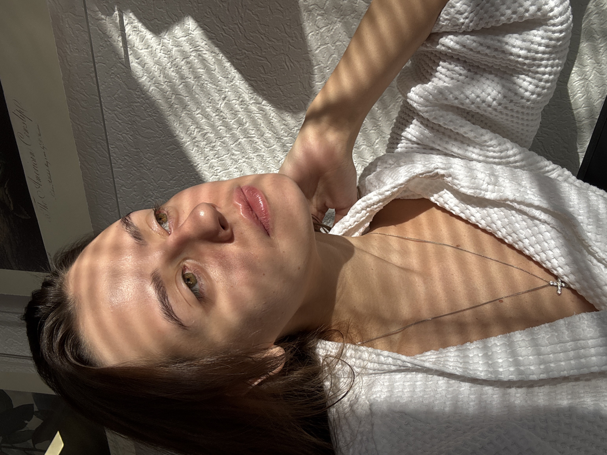 A person lies down in a white robe. The person is fair-skinned and has brown hair. There is a cross necklace on their neck. The light and shadow of window blinds are on the person's face and upper body. The background includes a wall with a textured surface. There is no evidence to suggest this image was taken in Moscow, Russia.