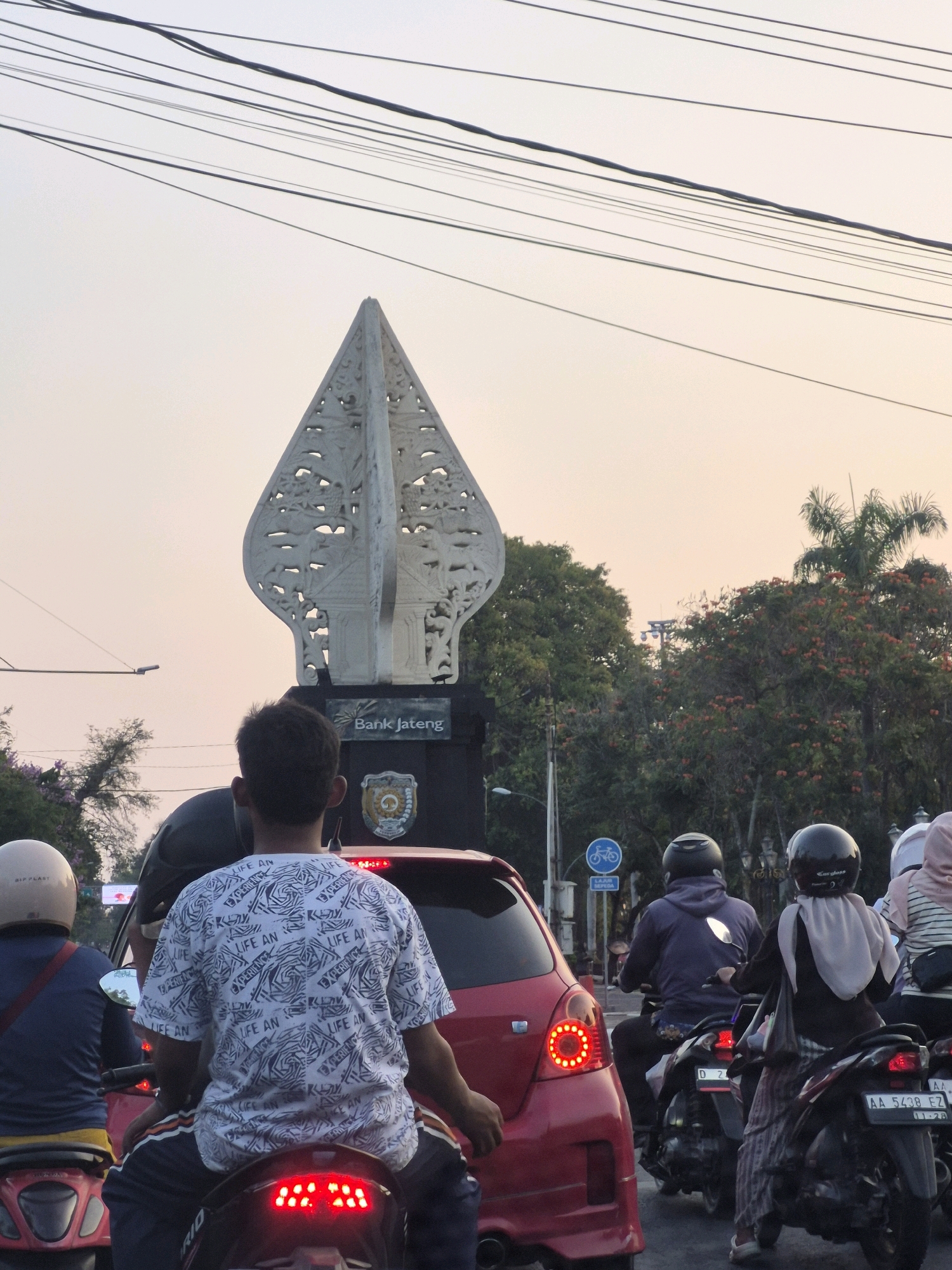 A daylight, medium shot captures a traffic scene in Purworejo, Indonesia. The frame is dominated by overhead power lines and a sky with a gradient of pale yellow and white. In the center background stands a large, ornate monument with a pointed top, resembling a Wayang Golek puppet's head, with intricate carvings. Below it, a sign reads "Bank Jateng". To the right, lush green trees with clusters of red flowers are visible, along with a tall palm tree.
In the foreground, several motorbikes and a red car are stopped, suggesting a traffic jam. On the closest motorbike, a person wearing a white t-shirt with a repeating black pattern of text and abstract shapes is seated, facing away from the camera. Another person on a motorbike to the left wears a blue shirt and a light-colored helmet. To the right, a person wearing a purple hoodie and a dark helmet is on a motorbike, followed by another person in a black hijab and a black helmet on a motorbike. A partially visible person in a white hijab is on the furthest motorbike on the right. The red car, positioned to the left of the central monument, has its taillights illuminated. The overall atmosphere is one of a bustling urban environment.