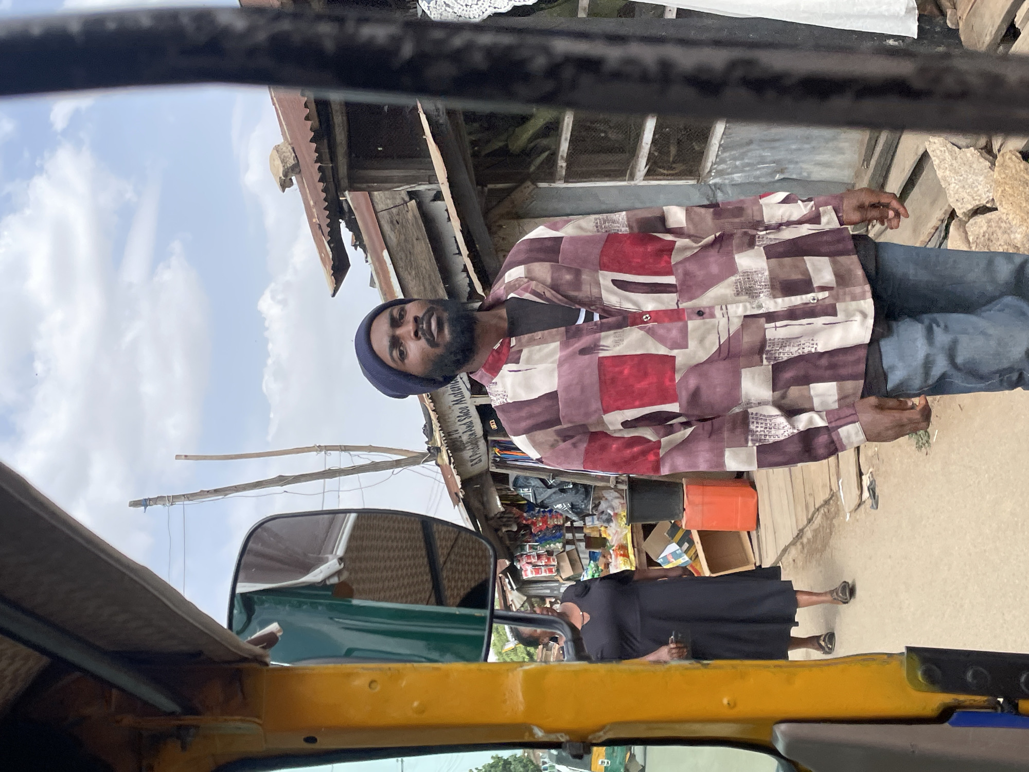 The image captures a street scene in Jos, Nigeria, likely from the perspective of someone inside a yellow tricycle taxi (keke napep). The photo is taken at eye level, looking across to the right side of the street.
In the foreground, part of the yellow vehicle and a green side mirror are visible, with a dark horizontal bar spanning the top of the frame, indicating the vehicle's interior structure.
The main subject is a man standing on the right side of the street, looking towards the camera. He has dark skin, a full beard, and is wearing a dark blue beanie. His shirt features a distinct patchwork design with squares of red, various shades of brown, and cream, worn over a dark undershirt. He is also wearing dark blue jeans. His expression is neutral.
Further in the background and to the right, a woman in a long black dress and dark sandals is visible, standing near what appears to be a stall or shop. She is holding a mobile phone in her left hand.
The background consists of a bustling street or market area with several makeshift structures. These buildings are constructed from corrugated metal sheets and wooden planks, suggesting commercial stalls or small shops. Various goods, including bottles, containers, and packaged items, are stacked on shelves and tables within these structures. A partially visible sign in the background reads "EJ Providing And Shoe Materials." The ground appears to be unpaved or dirt.
Above the structures, the sky is visible with scattered white clouds, indicating it is daytime with bright weather. Some wooden poles or beams are seen extending into the sky, possibly part of drying racks or support structures for awnings.