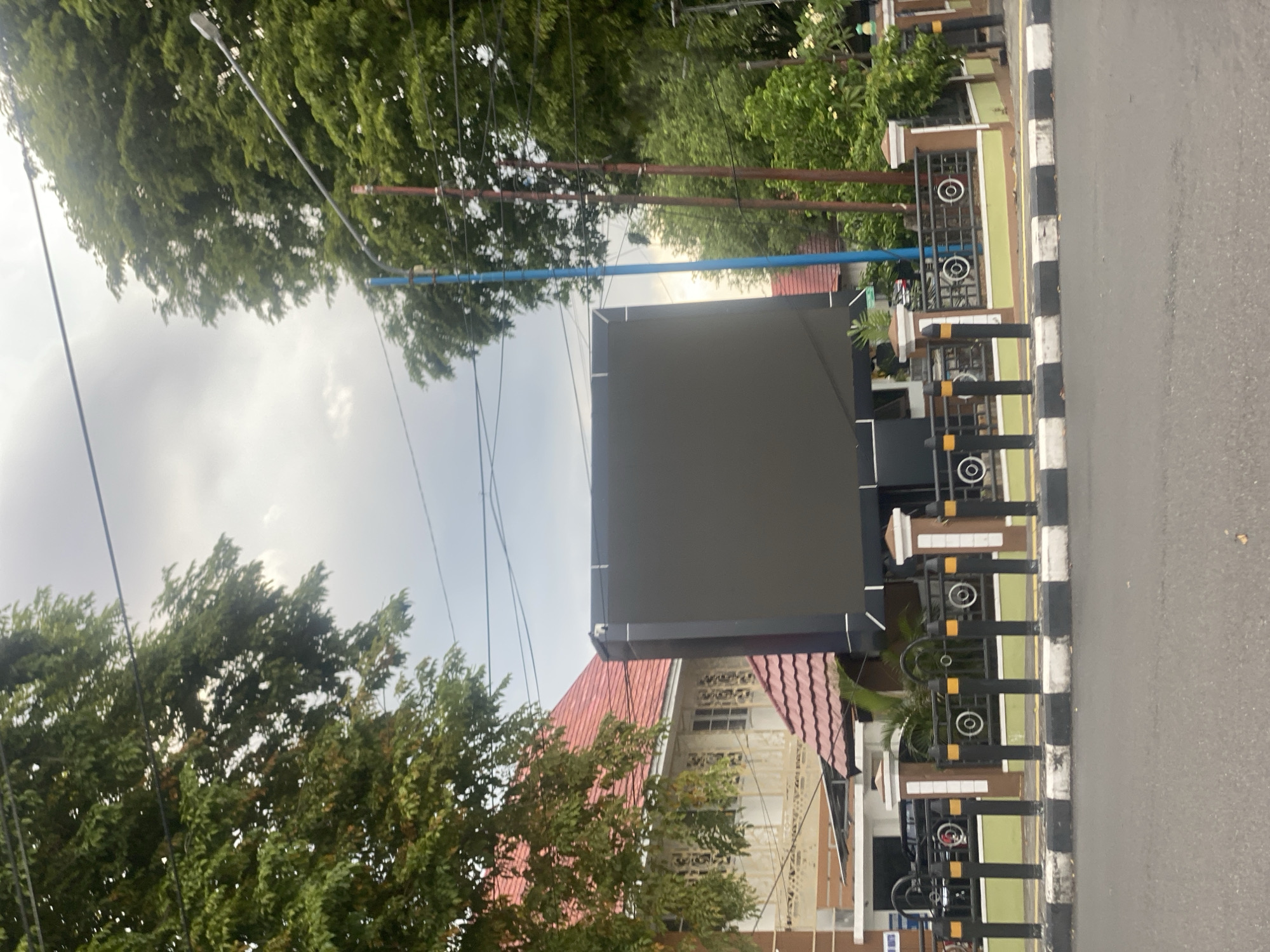 The image shows a large, dark, rectangular digital billboard in front of a building with a red tiled roof. The billboard is positioned on a street in Banjarbaru, Indonesia, with trees and power lines visible in the background. A decorative fence separates the billboard area from the road. The sky is overcast.