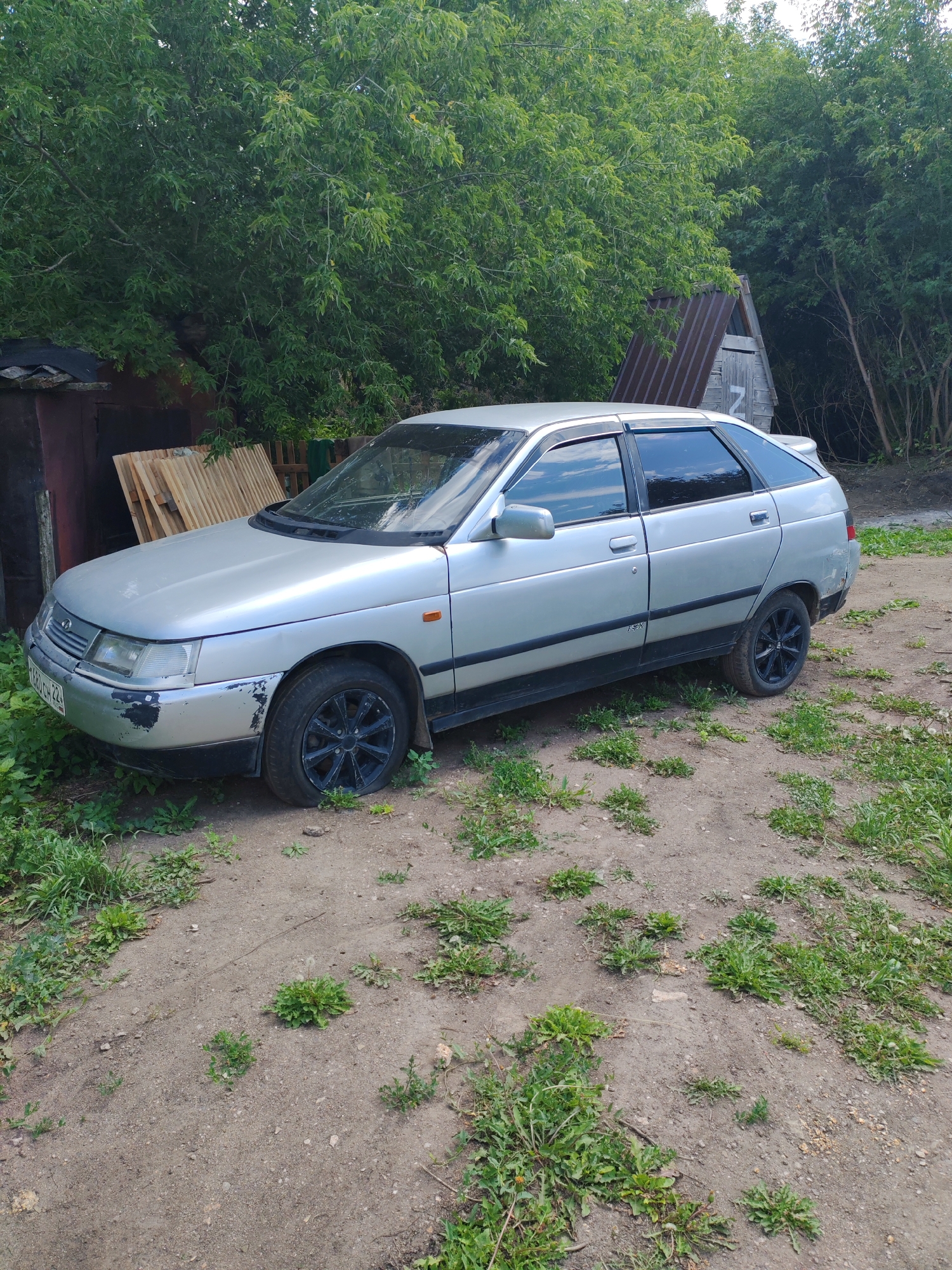 A silver hatchback car is parked on a dirt road surrounded by trees and vegetation. The car has tinted windows and black rims. The front bumper has visible damage and paint chips. To the left of the car, there is a lean-to structure with wooden planks stacked against it. In the background, a small, A-frame wooden structure with a corrugated metal roof is visible among the trees. The ground is a mixture of dirt and sparse green plants. The overall scene appears to be in a rural or semi-rural setting. The license plate of the car appears to read "K680CH 22".