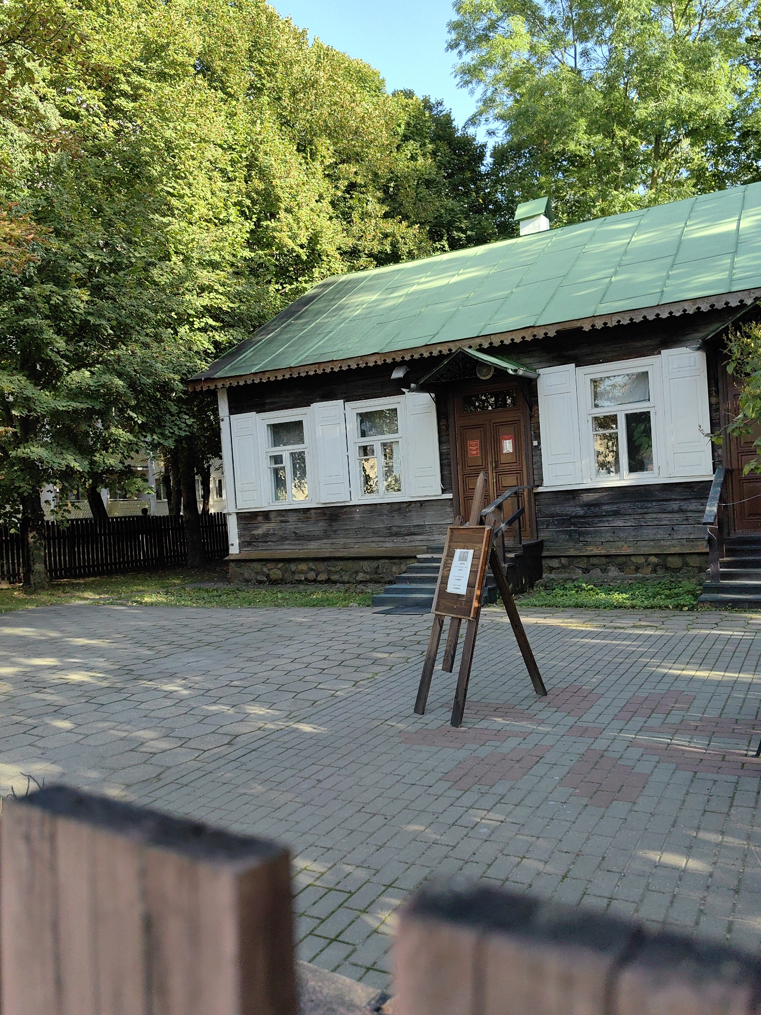 The image depicts a wooden house with a green roof, possibly a museum or historical building, located in Minsk, Belarus. The house has white shutters on its windows and decorative wooden trim under the eaves. In front of the house, a paved courtyard is visible, with a wooden easel displaying a sign. Tall green trees with lush foliage surround the building and the courtyard. The sky is clear and blue, suggesting it is daytime. A low wooden fence is partially visible in the background. The scene appears to be calm and quiet, with no people actively engaged in any visible activity. The overall impression is one of a serene and well-maintained historical site.