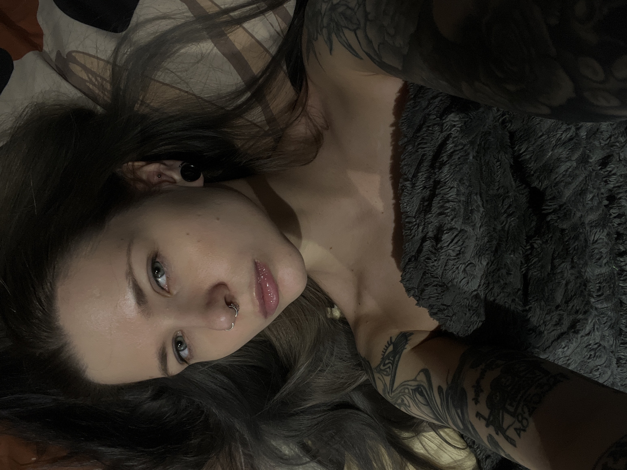 An eye-level indoor portrait shows a person, likely a young woman, lying down and looking directly at the camera. The setting appears to be a dimly lit room, possibly a bedroom in Khimki, Russia, with soft furnishings including a patterned pillow and a dark grey, plush, textured blanket.
The subject has long, dark hair spread around her head and shoulders. Her light-colored eyes (possibly blue or green) are focused on the viewer, and her expression is neutral and calm. She wears a silver hoop nose piercing and a dark ear tunnel/gauge along with a small stud in her visible ear. Her lips are full and appear to be covered in gloss. Visible tattoos cover her arms and extend towards her shoulder and neck area, featuring dark, intricate designs. Some partially legible text, possibly including "LAB", can be seen within a tattoo on her right forearm. She is partially covered by the plush blanket, which drapes over her torso and arms. The overall lighting is soft and subdued.