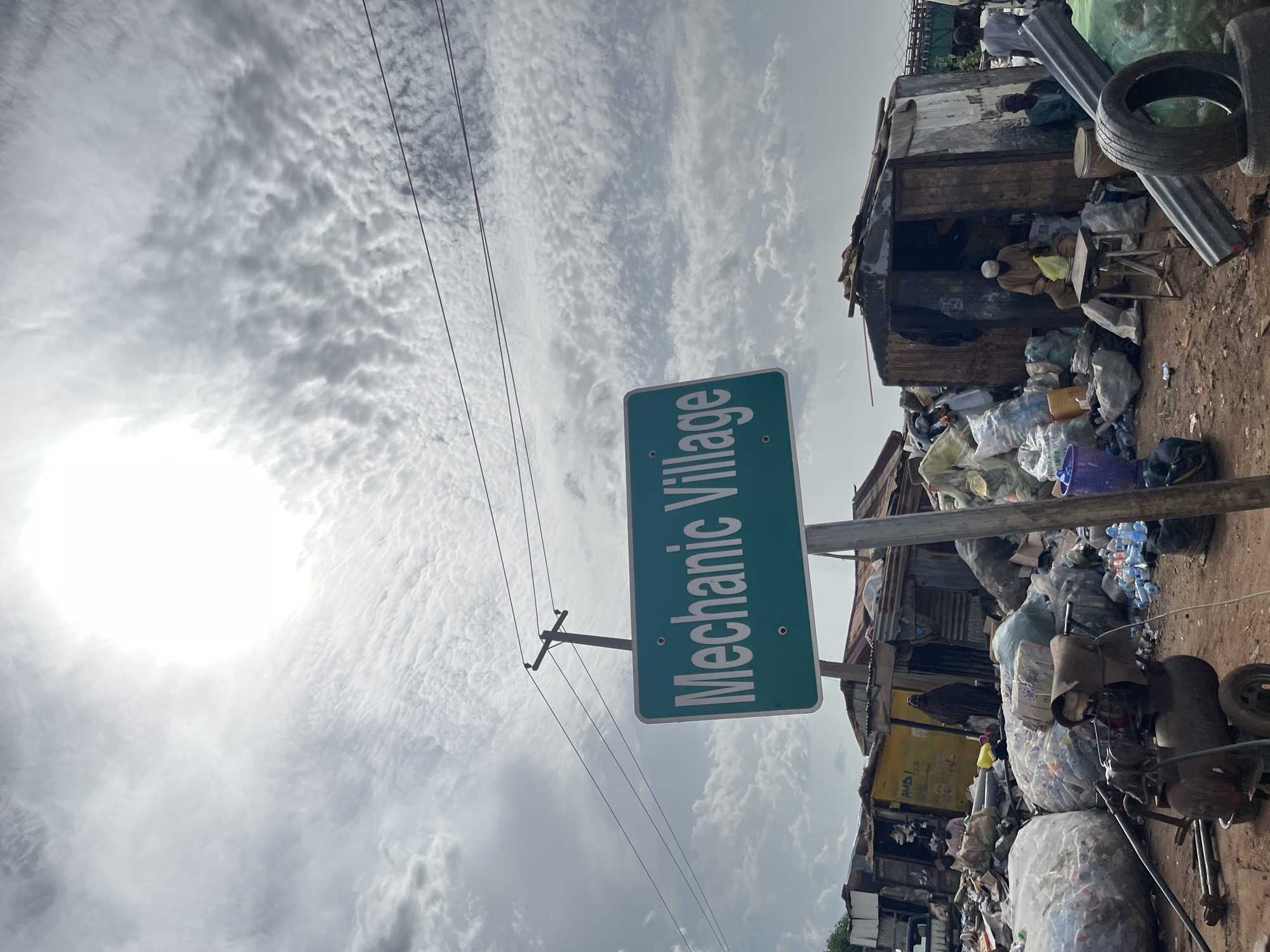The image captures a scene in Jos, Nigeria, depicting the "Mechanic Village." The overall atmosphere appears to be one of informal commerce and resourcefulness amidst a setting that is heavily laden with various objects, predominantly waste and salvaged materials.
The primary subject is the sign itself, a green rectangular sign with white lettering that reads "Mechanic Village." This sign is mounted on a pole and is positioned vertically in the frame due to the image's orientation. Surrounding the sign is a bustling environment characterized by piles of what appear to be recyclable materials, including plastic bottles, bags of refuse, and discarded tires.
In the background, there are structures that seem to be makeshift stalls or workshops, constructed from corrugated metal and wood. People are visible within these structures and around them. One person, wearing a light-colored garment and a face covering, is seated at a small table in front of one of the stalls, appearing to engage in some activity. Other individuals are also present in the background, suggesting ongoing work or trade.
The scene is set under a cloudy sky with the sun partially obscured, indicating daytime, likely mid-morning or afternoon. The lighting suggests an overcast or partly sunny condition. The ground is dirt and appears uneven, consistent with an outdoor, less developed area. The sheer volume of discarded items points to a recycling or scrap yard operation within the mechanic village. The overall impression is one of a working community that thrives on the repurposing of materials. There is no explicit indication of emotions, but the scene suggests resilience and an active livelihood.