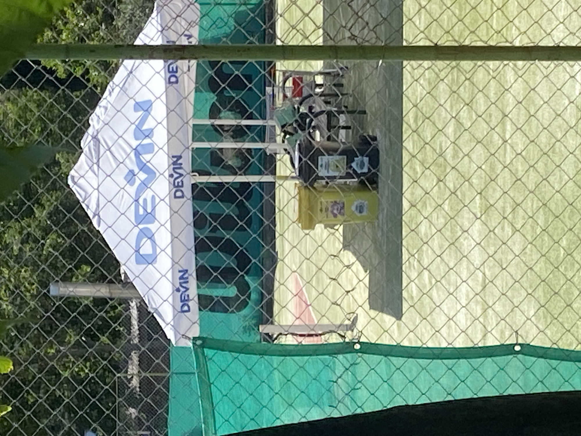 The image shows a tennis court with a large white tent canopy providing shade. The canopy has the word "DEVIN" printed on it in blue letters. In front of the tent, there are two trash bins, one yellow and one dark gray. Behind the tent, there are chairs and tables. The tennis court is fenced with a metal chain-link fence covered in parts with green netting. Trees are visible in the background. The scene appears to be outdoors in a public area.