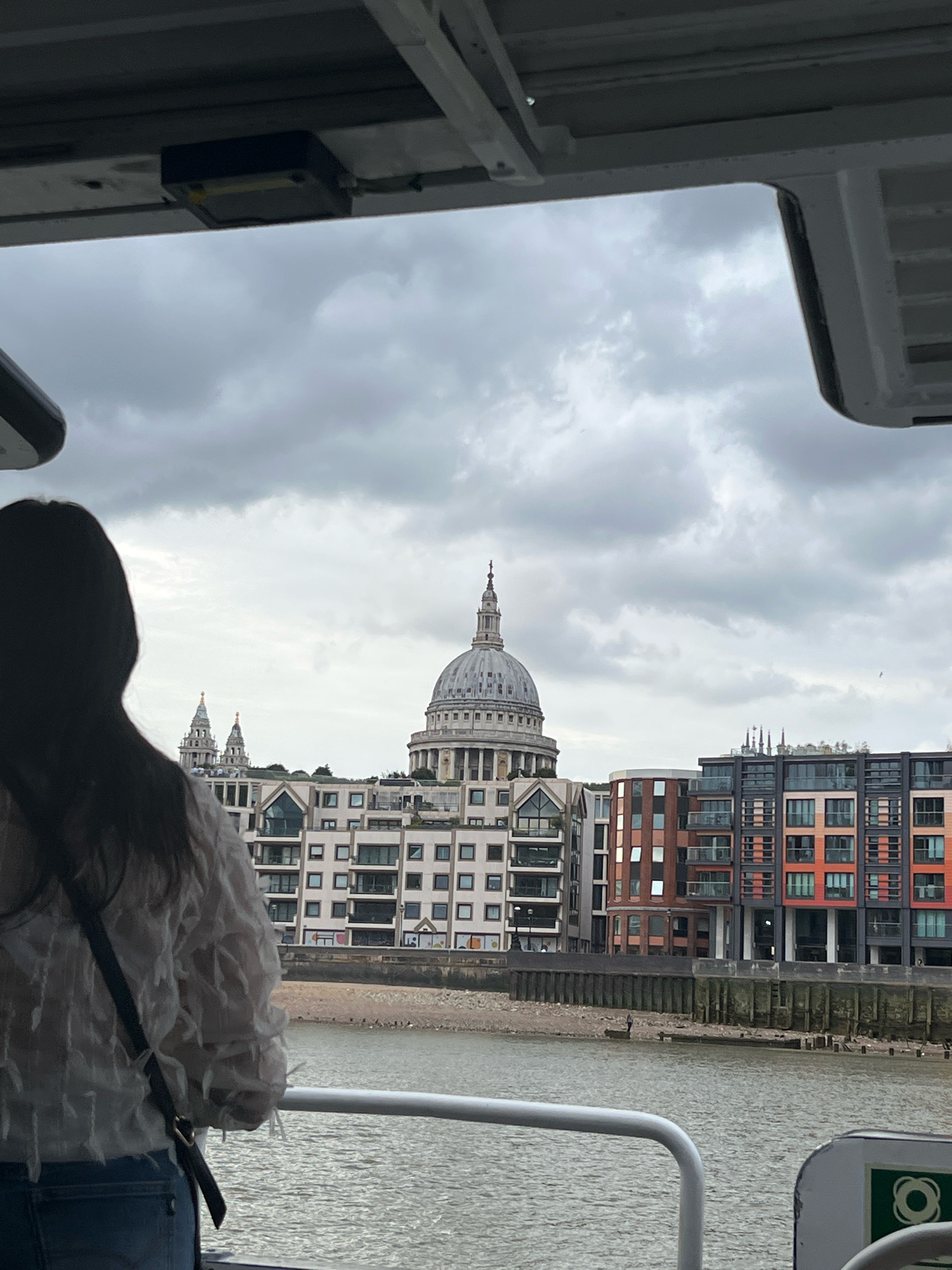 The image is a view of the River Thames in London, United Kingdom, taken from what appears to be a boat or a ferry. In the foreground, the back of a woman wearing a white, textured blouse and blue jeans is visible. She is looking out at the scene. The river occupies the lower part of the image, with a light railing of the boat in the immediate foreground. Beyond the river, the London skyline is visible. Dominating the center of the skyline is the iconic dome of St. Paul's Cathedral. To the left of St. Paul's, two smaller church spires can be seen. In front of the cathedral and spires, a row of modern apartment buildings with a mix of white, grey, and colorful facades runs along the waterfront. A concrete embankment and a gravelly shore separate the river from the buildings. The sky is overcast with heavy clouds, suggesting a typical London day.