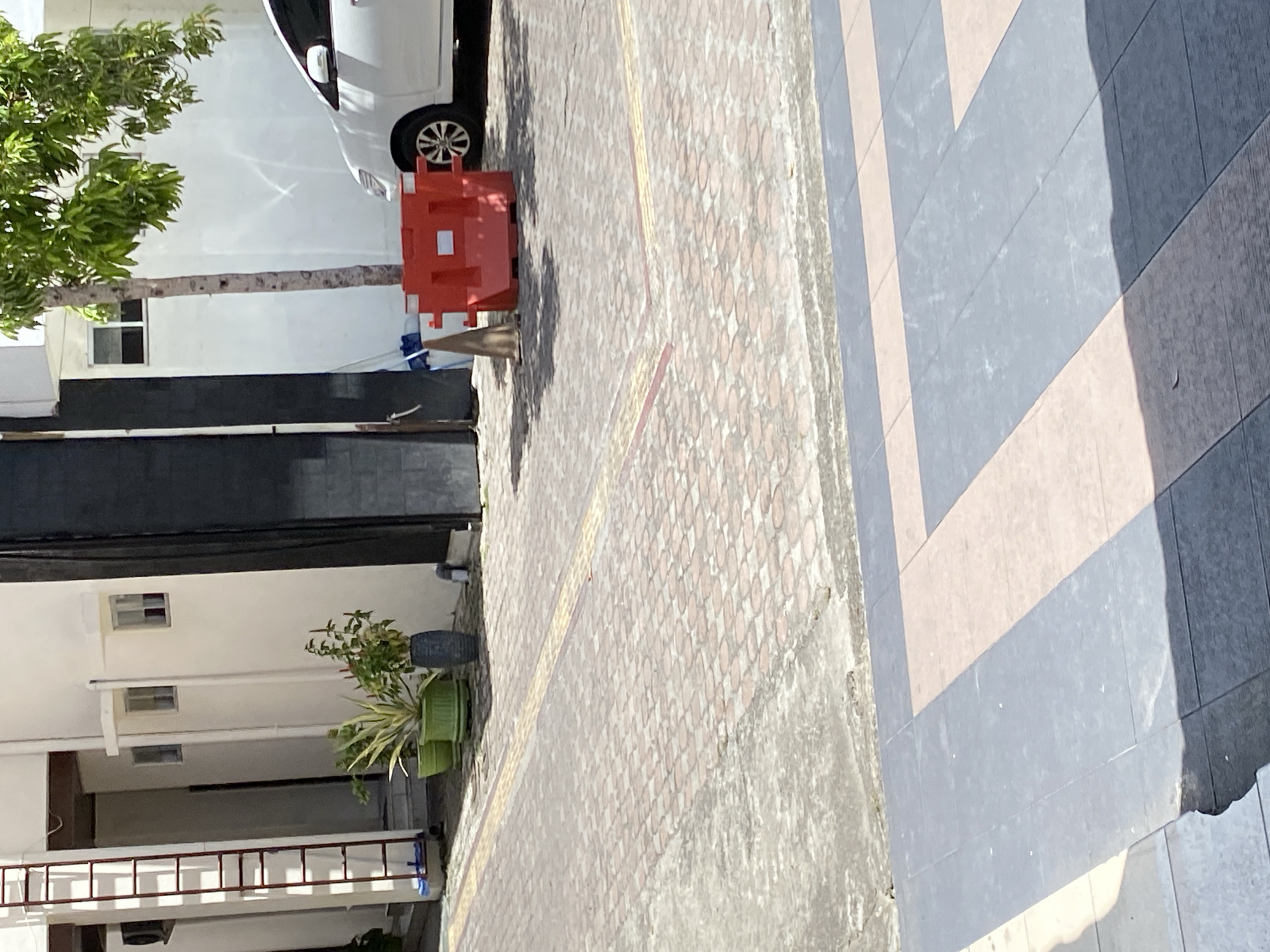 The image shows a paved area in front of a building, likely in Banda Aceh, Indonesia. A white car is parked on the right side of the frame. In the foreground, there are large, dark gray and light brown tiles laid out in a pattern. The midground features a patterned brick-like pavement with yellow tactile paving strips. Several potted plants, including a variegated leaf plant in a green pot, are situated near the building's wall. A red plastic barrier is placed on the pavement. A tall, thin tree grows to the right of the building. A red ladder is leaning against the left side of the building. The building itself is white with some dark paneling. The scene appears to be an outdoor space associated with a commercial or residential property.